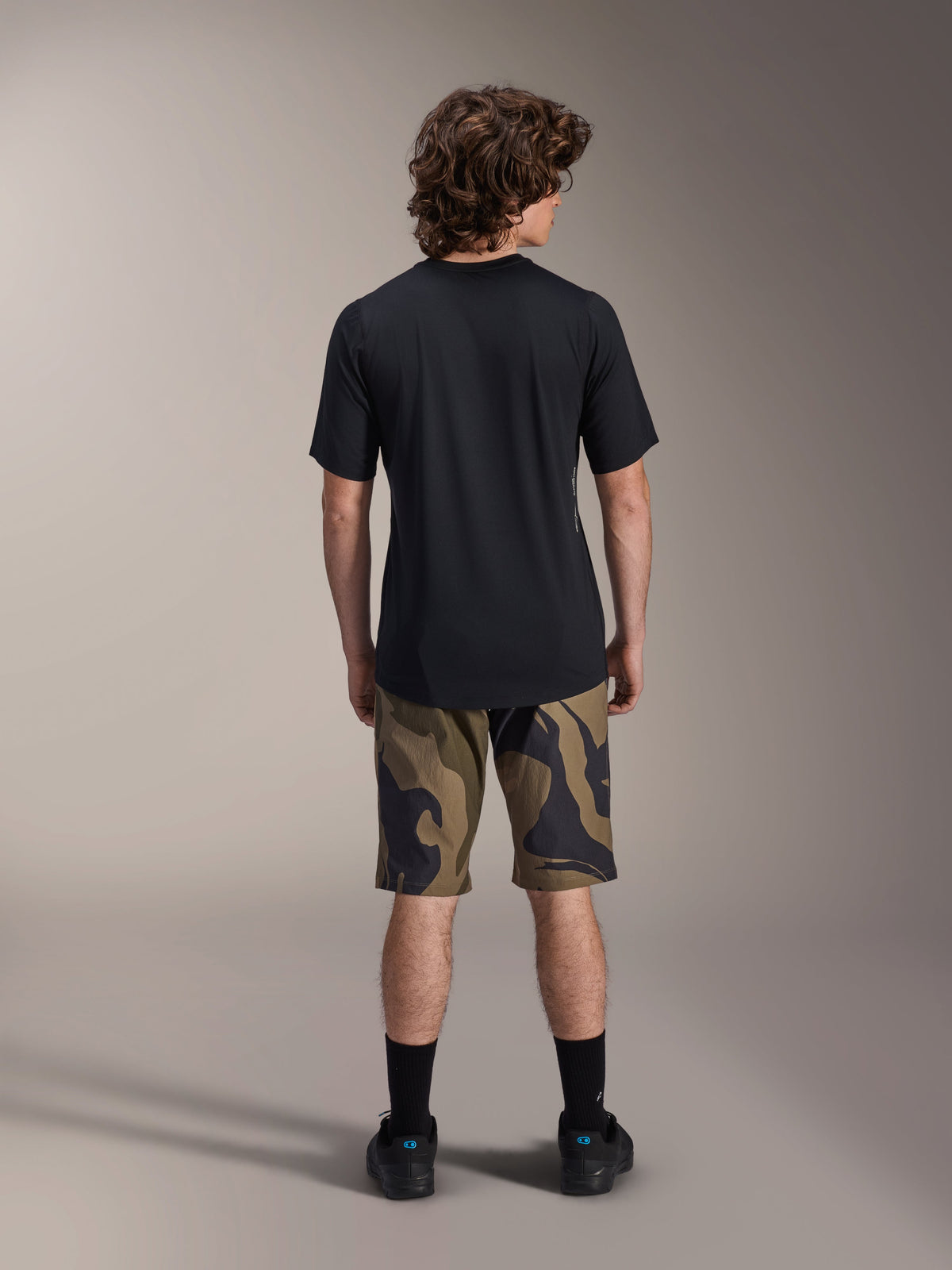 Alpinestars A-Dura Astar Kurzarm-Trikot, Schwarz, Rückansicht an einem Modell, kombiniert mit MTB-Shorts mit Camouflage-Muster und schwarzem Schuhwerk, lässige technische Mountainbike-Bekleidung