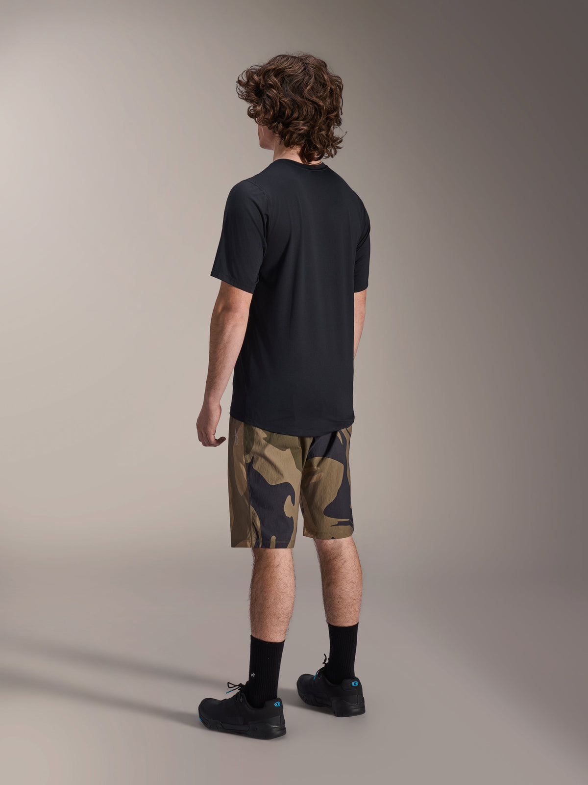 Alpinestars A-Dura Astar Kurzarm-Trikot in Schwarz, Rückansicht, kombiniert mit Shorts im Camouflage-Muster sowie schwarzen Sportsocken und Schuhen, lässige MTB-Ausrüstung