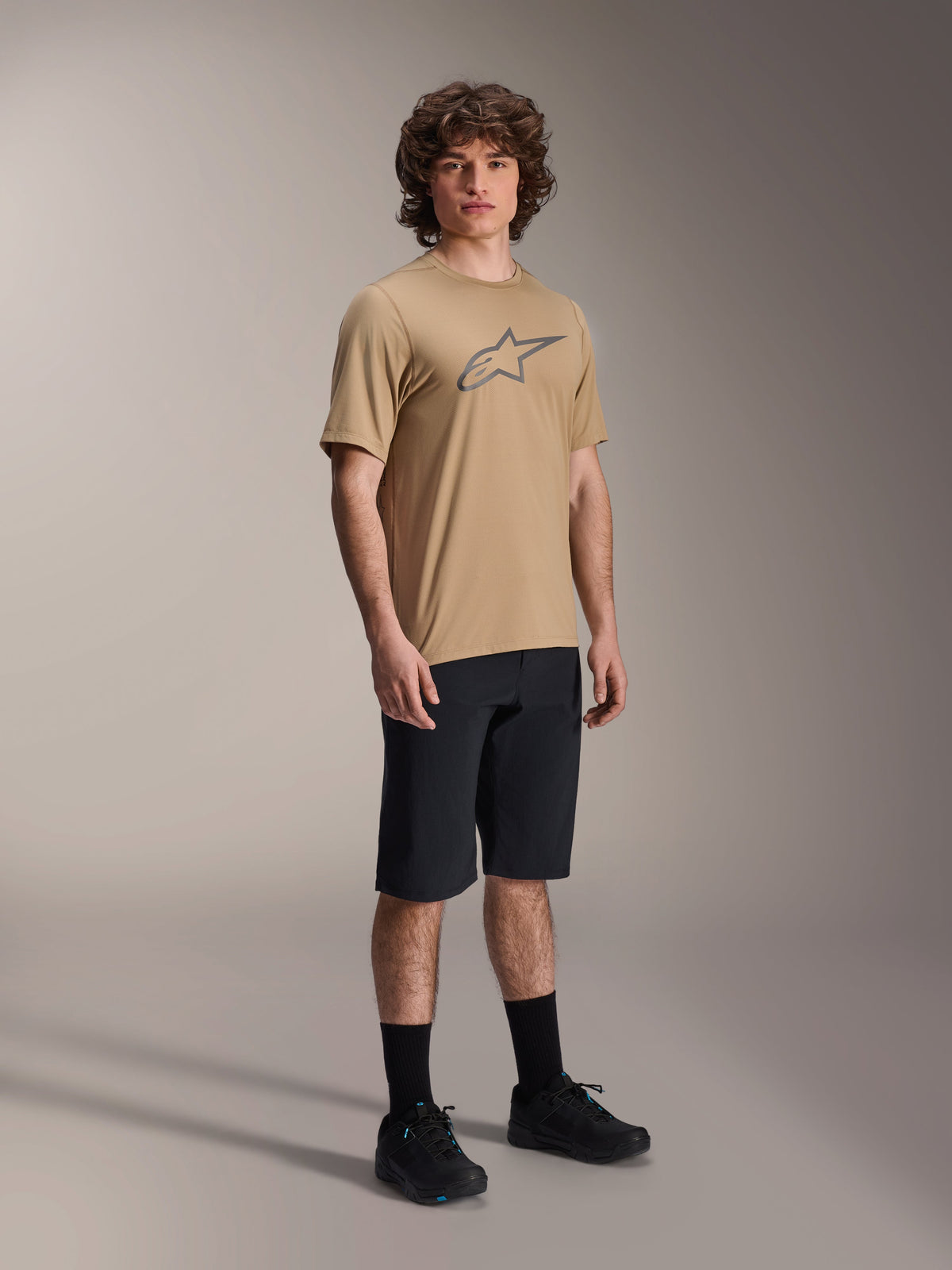 Alpinestars A-Dura Astar Short Sleeve Jersey, Light Brown, featuring a large dark gray Astar logo on the chest, worn by a model with black MTB shorts, black socks and black shoes, casual performance mountain bike apparel in a studio setting