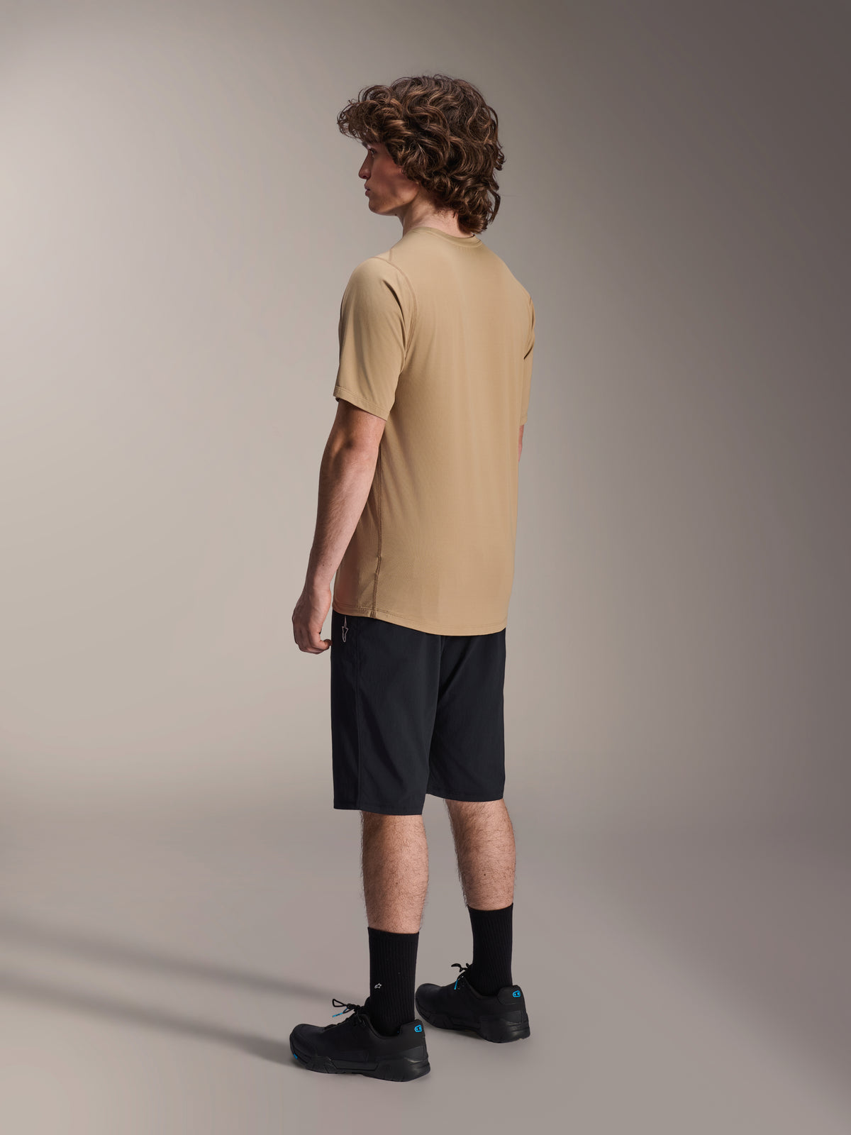 Alpinestars A-Dura Shorts, Mountainbike-Shorts, Schwarz, kombiniert mit einem sandfarbenen kurzärmeligen A-Dura Trikot und schwarzen Radschuhen, mit regulärer Passform und strapazierfähiger Konstruktion für den Offroad-Radsport, seitliche Rückansicht