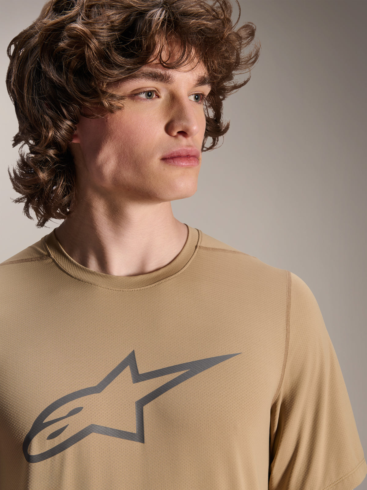 Model wearing Alpinestars A-Dura Astar Short Sleeve Jersey, Light Brown MTB Jersey, featuring a grey star logo on the chest, moisture-wicking jacquard fabric, casual cycling style