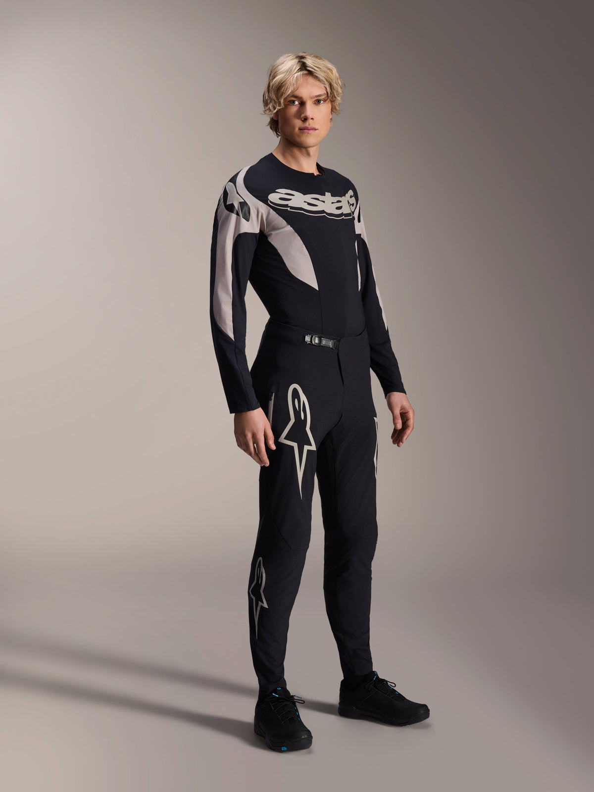 Male model wearing Alpinestars A-Supra Melt Pants and matching Long Sleeve Jersey, Black with light grey curved patterns, featuring bold white Astars logos on the legs and wordmark on the chest, professional mountain bike racing outfit.