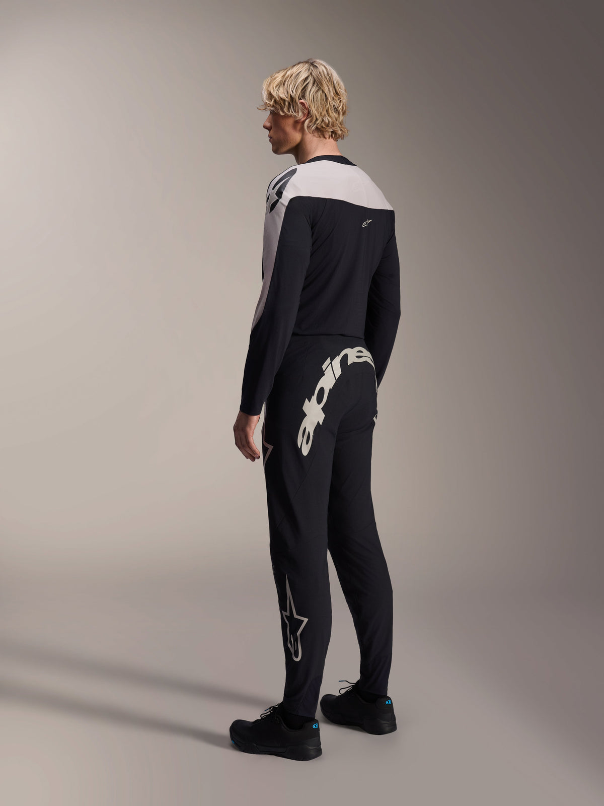 Alpinestars A-Supra Melt Pants, MTB Pants, Black, featuring a large white Alpinestars wordmark on the back yoke and star logo on the leg, worn by a model with a matching long-sleeve jersey and black shoes, back-side view