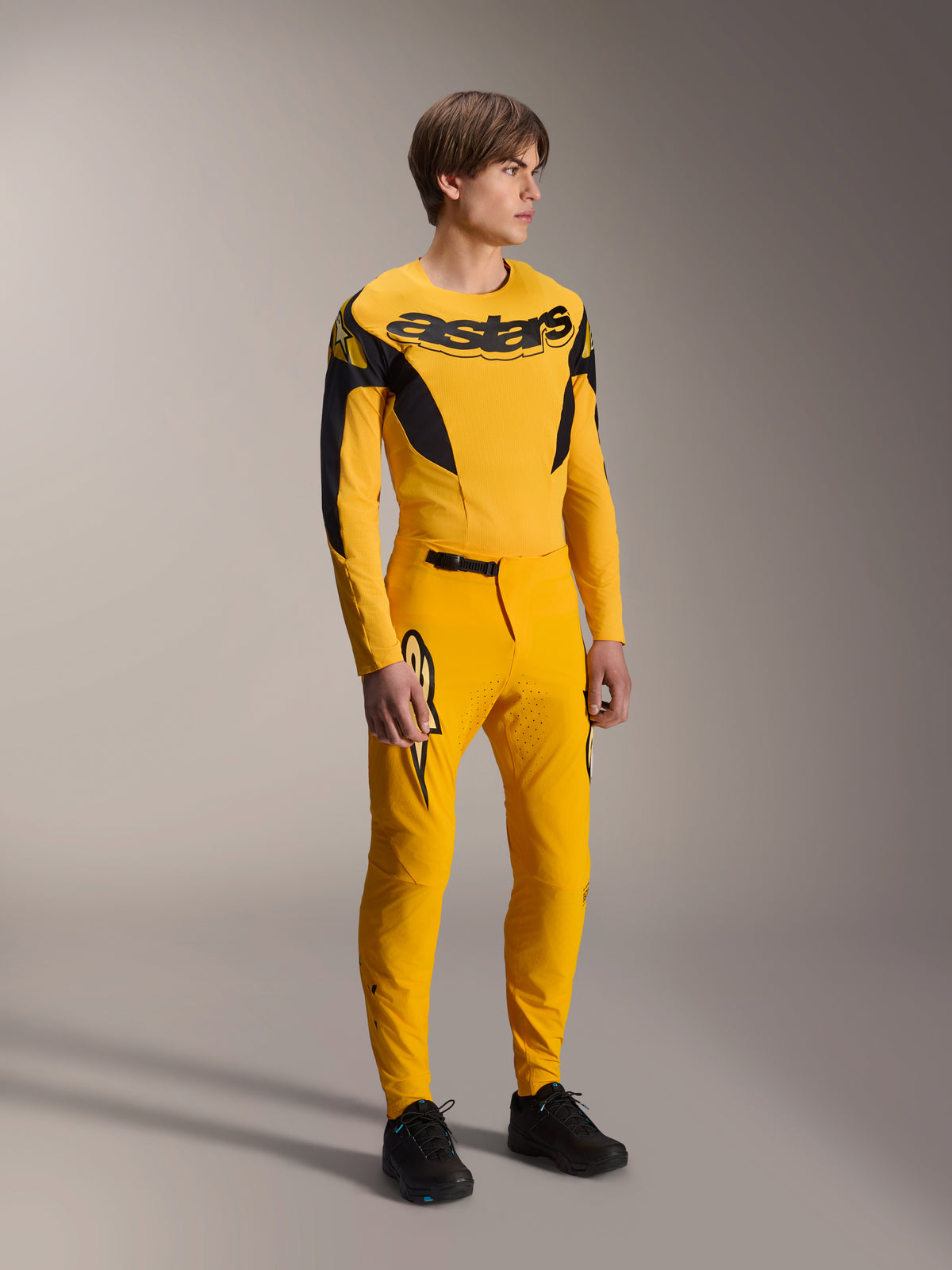 Männliches Model im Alpinestars Limited Edition A-Supra Melt Gold Gear-Set, gelbes langärmeliges MTB-Trikot mit markantem schwarzem Branding auf der Brust, passende gelbe Hose mit lasergeschnittener Belüftung und schwarzen Akzenten, komplette Mountainbike-Rennausrüstung.