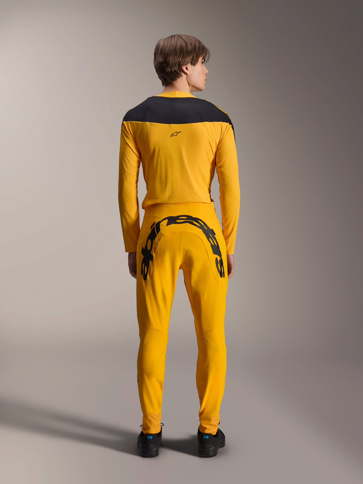 Rückansicht eines Modells im Alpinestars Limited Edition A-Supra Melt Gold Gear-Set, bestehend aus einem dunkelgelben Langarmtrikot mit schwarzer Schulterpasse und einer passenden dunkelgelben Hose mit einem großen schwarzen Alpinestars-Schriftzug auf der Rückenpasse, kombiniert mit schwarzen Schuhen.