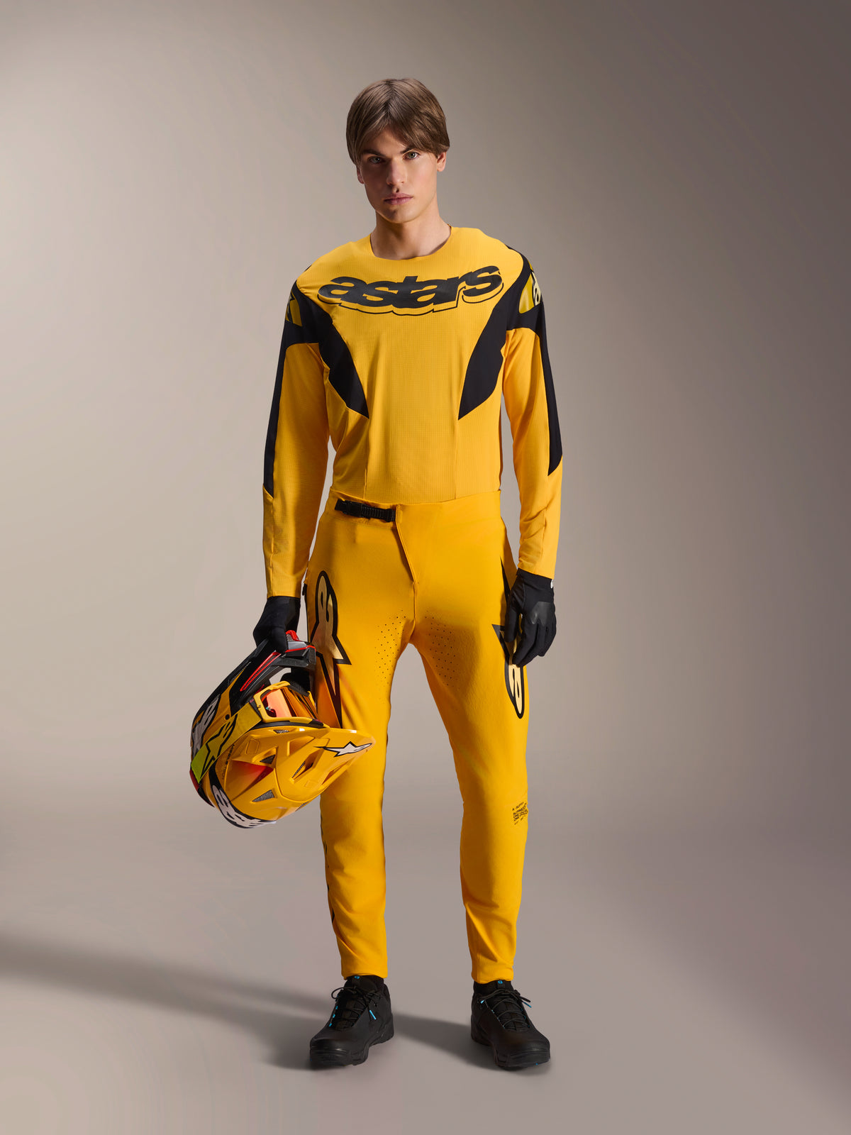 Model trägt Alpinestars Limited Edition A-Supra Melt Gold MTB-Kit, gelbes Langarmtrikot mit schwarzem „astars“-Logo, passende gelbe Hose mit schwarzen Akzenten, hält einen gelb-schwarzen Rennhelm, schwarze Handschuhe und Schuhe, Performance-Ausrüstung für Mountainbiking