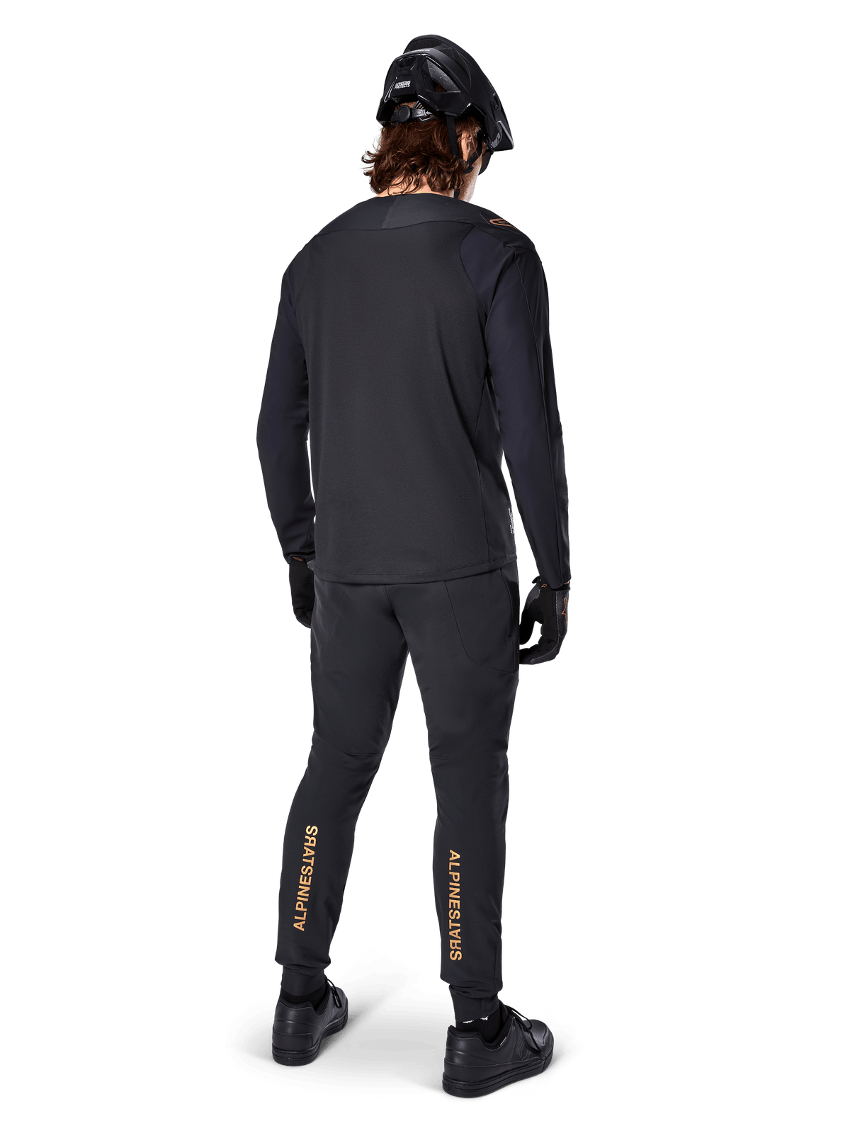 Rückansicht eines Mountainbike-Fahrers im Alpinestars A-Aria Polartec Switch Langarmtrikot in Schwarz, kombiniert mit einer schwarzen MTB-Hose mit hellbraunem Alpinestars-Branding an den Waden, schwarzem Helm und Handschuhen, professionelle Radsportausrüstung für Offroad-Trails.