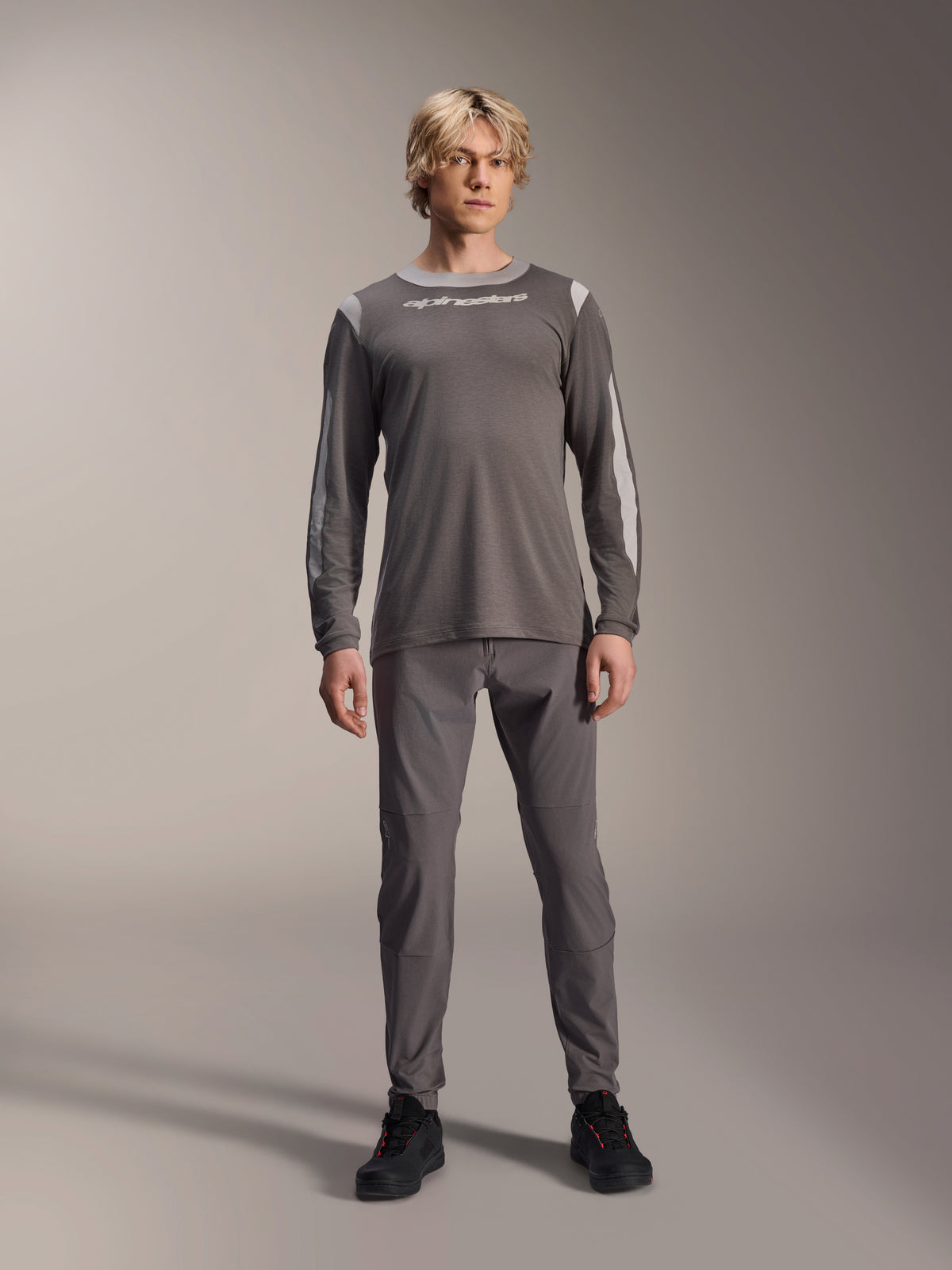 Alpinestars A-Dura Dri Wool Langarmtrikot, Dunkelgrau, getragen von einem Modell mit passender grauer Hose und schwarzen Schuhen, weißes Alpinestars-Branding auf der Brust, hellgraue Mesh-Einsätze an Schultern und Ärmeln, technische Mountainbike-Ausrüstung
