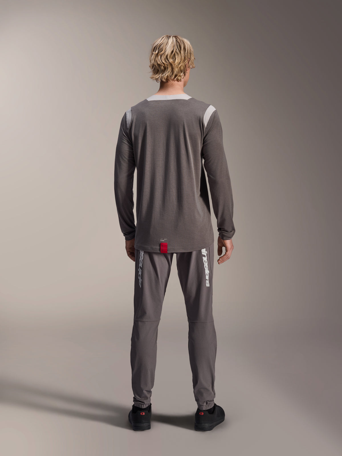 Alpinestars A-Dura Dri Wool Langarmtrikot, Dunkelgrau, Rückansicht am Modell, mit hellgrauen Mesh-Einsätzen an Schultern und Kragen, verlängerter Rückensaum mit kleinem roten Markenlabel, getragen mit passender grauer Radhose.