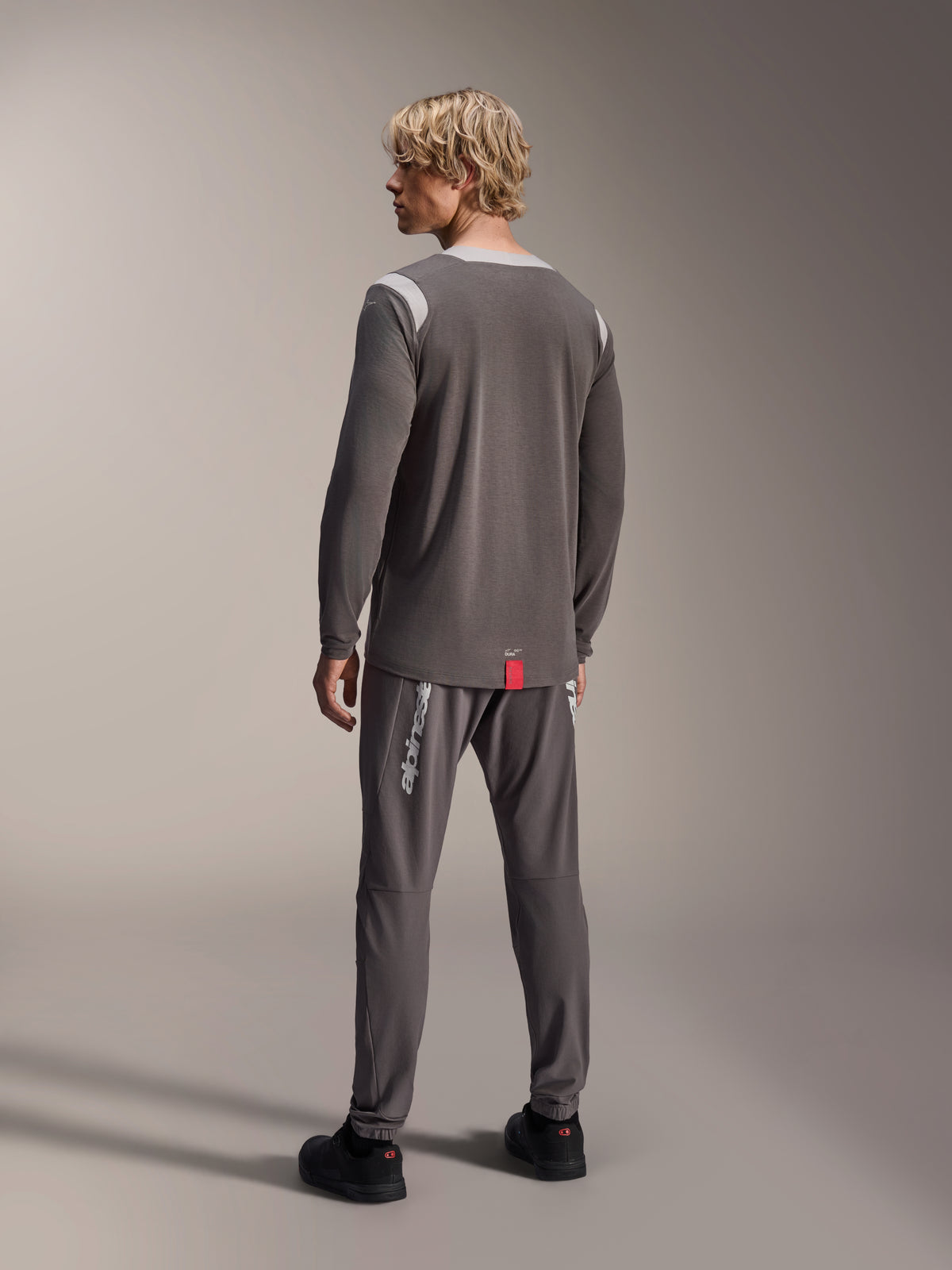 Männliches Model im Alpinestars A-Dura Dri Wool Langarmtrikot, Dunkelgrau mit hellgrauen Mesh-Schultereinsätzen, kombiniert mit einer passenden dunkelgrauen Radhose, Rückansicht zeigt den verlängerten Rückensaum, Mountainbike-Bekleidung
