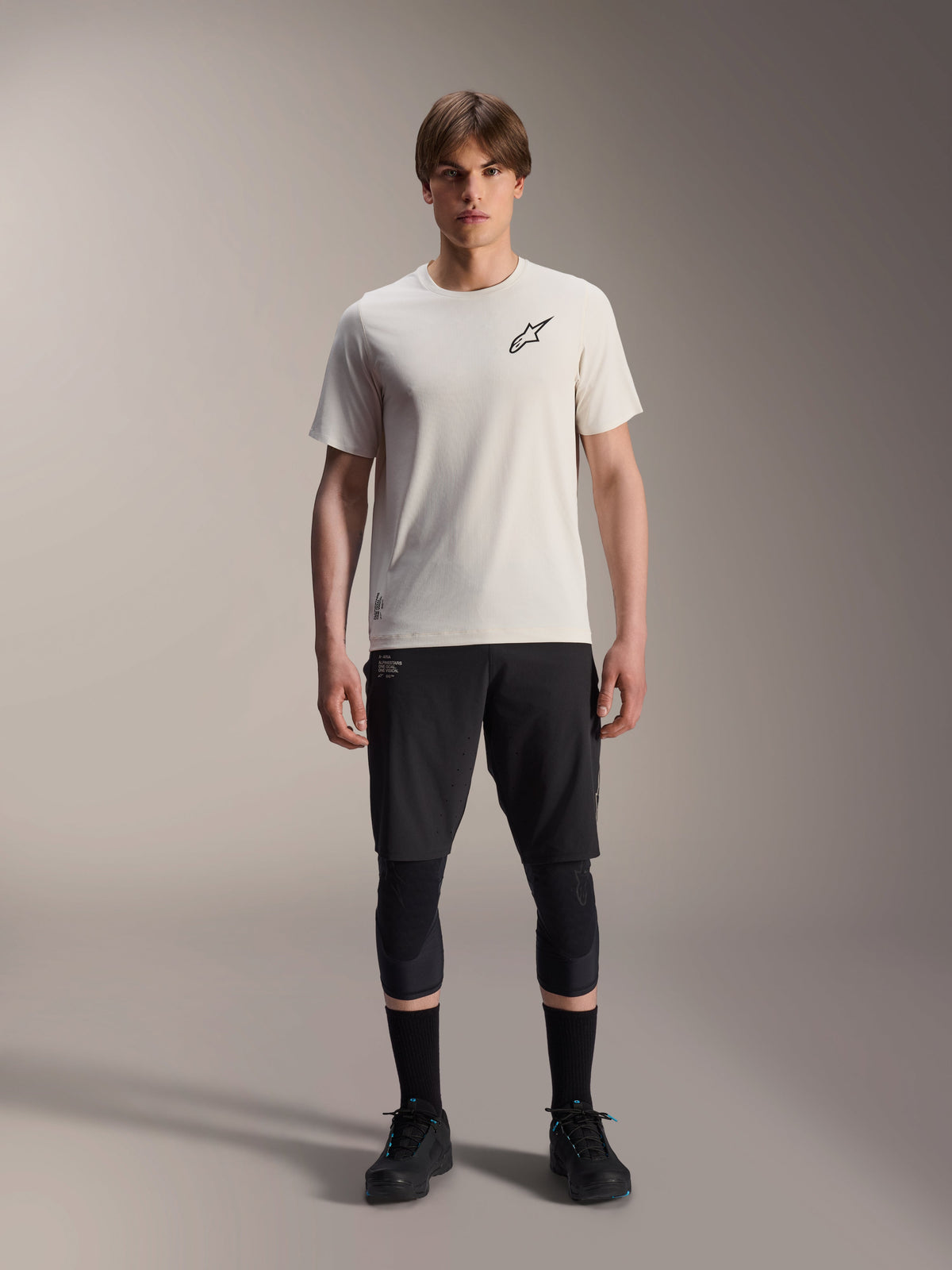 Männliches Model trägt das Alpinestars A-Aria Astar Kurzarmtrikot in Ecru, kombiniert mit schwarzen MTB-Shorts, Knieschonern und schwarzen Radschuhen, mit einem minimalistischen schwarzen Stern-Logo auf der Brust, konzipiert für Offroad-Trailfahrten.