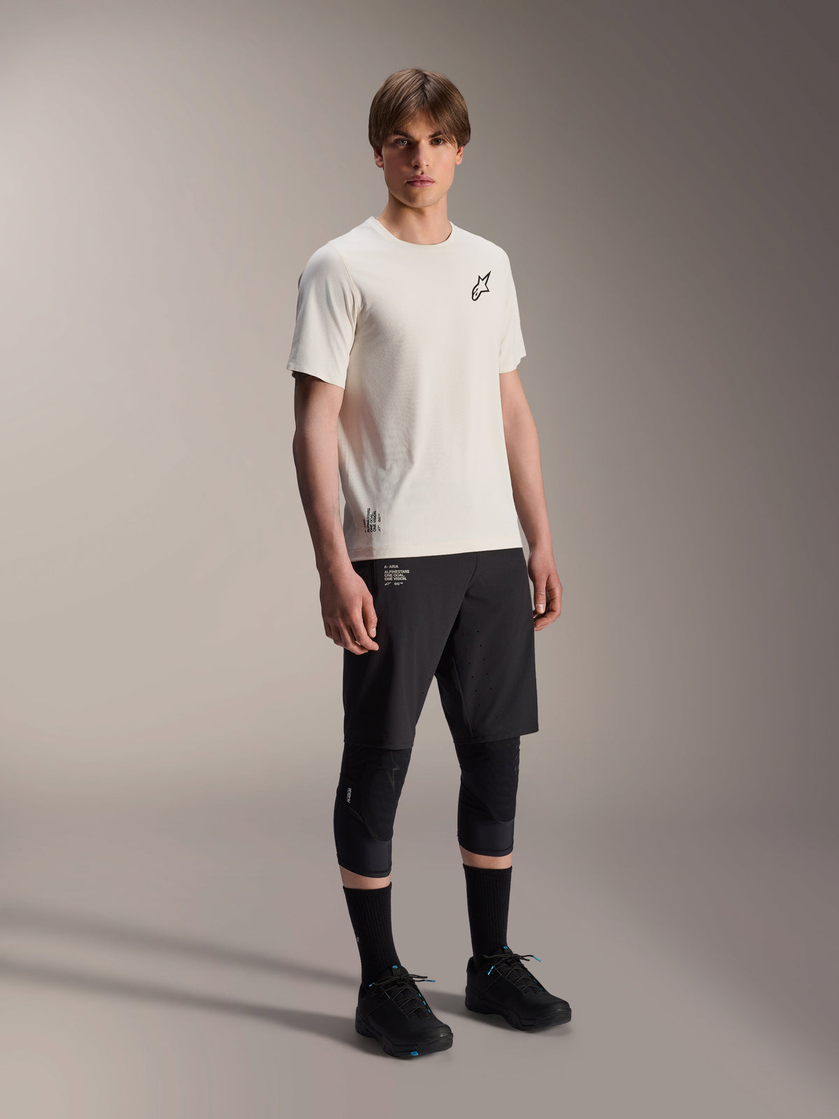 Alpinestars A-Aria Astar Kurzarmtrikot, Ecru, mit schwarzem Brust-Logo, getragen mit schwarzen Radshorts und Knieprotektoren, leichte MTB-Performance-Ausrüstung
