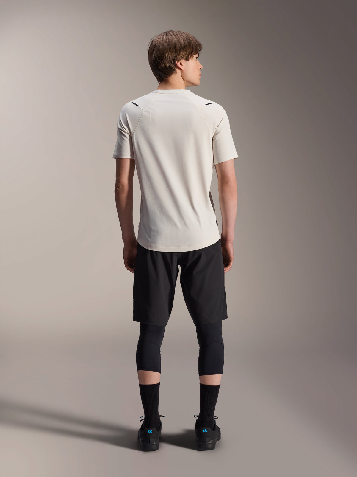 Rückansicht eines männlichen Models im Alpinestars A-Aria Astar Kurzarmtrikot in Ecru, kombiniert mit schwarzen MTB-Shorts und Schutz-Leggings, verlängerte Rückenpartie, Performance-Passform für Offroad-Trailfahrten.