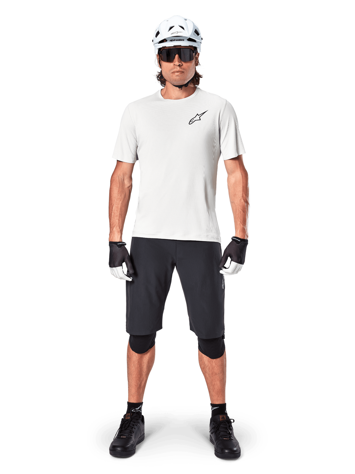 Männlicher Radfahrer in Alpinestars A-Aria Astar Kurzarmtrikot in White Dove, mit schwarzem Astars-Logo auf der Brust, kombiniert mit schwarzen Mountainbike-Shorts, weißem Helm, Sonnenbrille, schwarzen Knieprotektoren und Handschuhen, komplettes Offroad-Radsport-Outfit