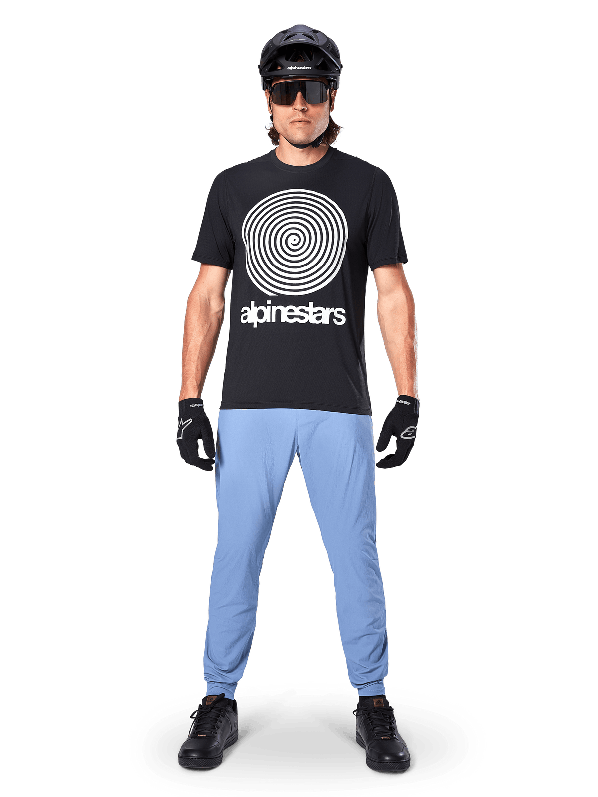 Alpinestars A-Dura Oscar Kurzarmtrikot, schwarzes MTB-Trikot, mit einer großen weißen Vintage-Oscar-Spiralgrafik und Markenschriftzug, getragen mit hellblauer Hose, schwarzem Helm, Sonnenbrille und schwarzen Handschuhen, Performance-Mountainbike-Bekleidung