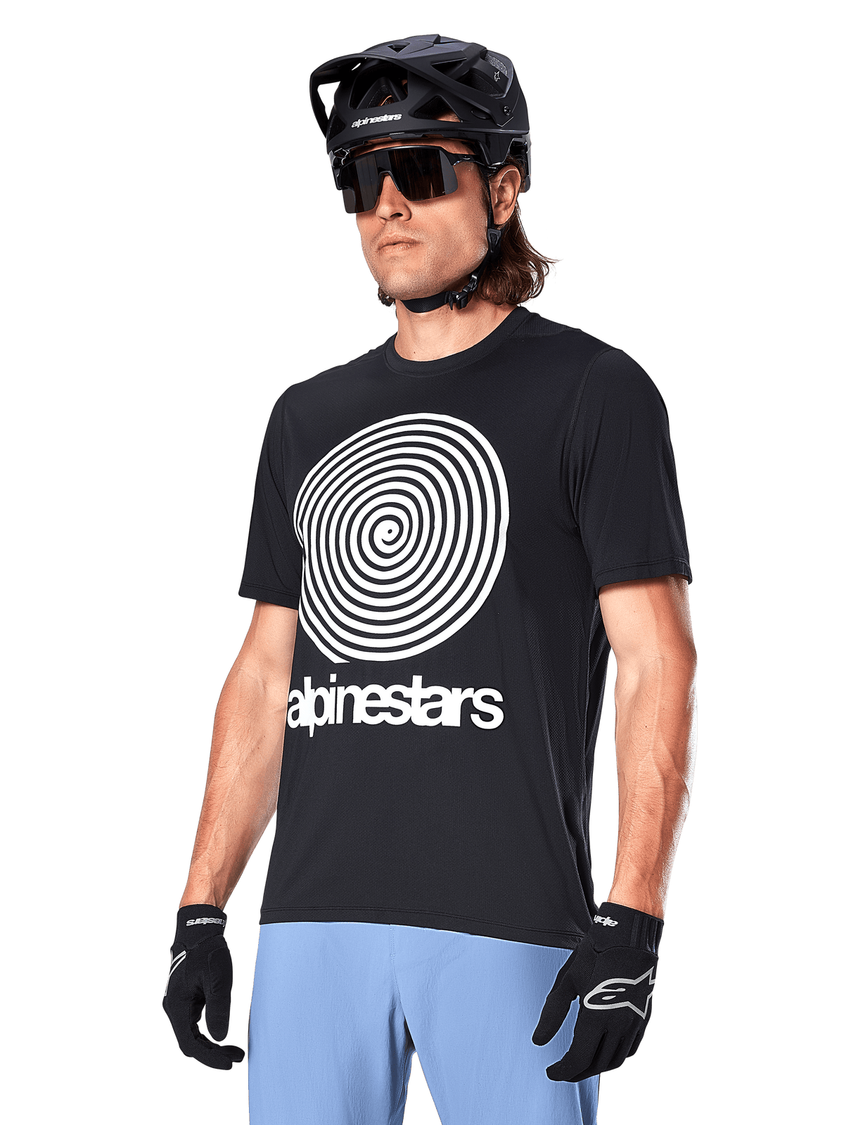 Alpinestars A-Dura Oscar Kurzarmtrikot, Schwarz, mit großer weißer Spiralgrafik und Alpinestars-Schriftzug auf der Brust, getragen von einem Fahrer mit schwarzem MTB-Helm, Sonnenbrille und hellblauer Hose
