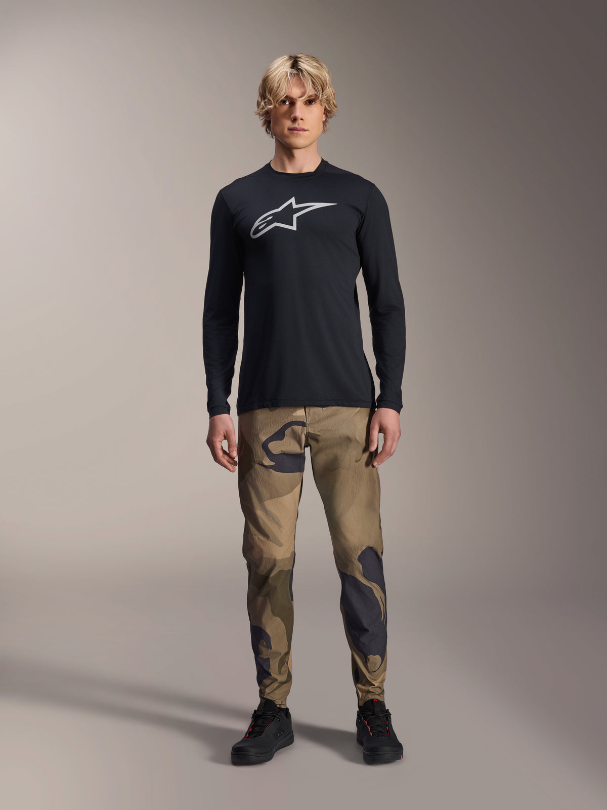 Alpinestars A-Dura Astar Long Sleeve Jersey, Black, featuring a large white Astars logo on the chest, worn with camouflage pants and black shoes, casual mountain bike performance wear