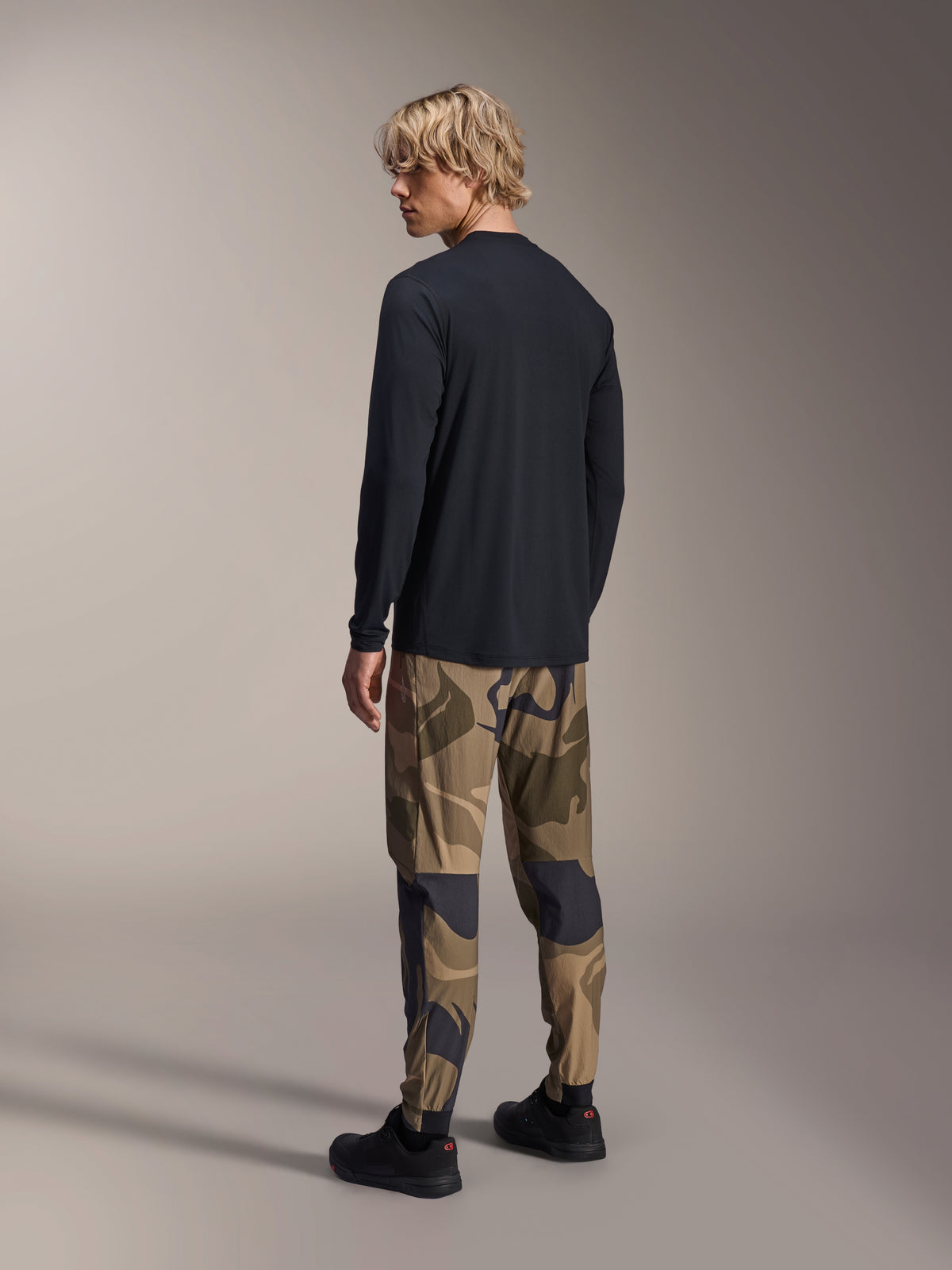 Alpinestars A-Dura Camo Pant, MTB Pants, Brown Camouflage, worn with a black long-sleeve jersey and black sneakers, rear view showing tapered legs and elasticated cuffs, performance apparel for off-road cycling
