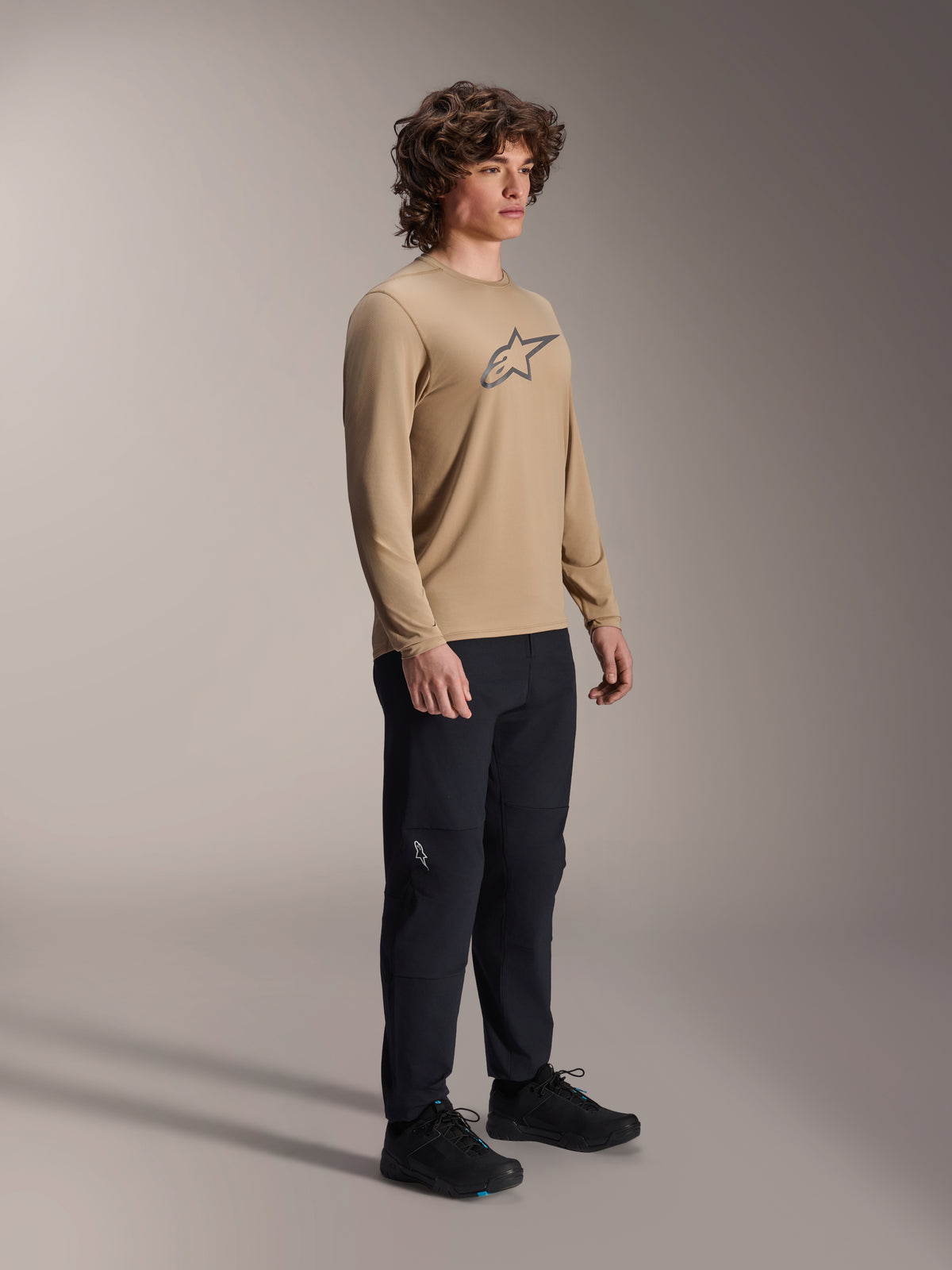 Alpinestars A-Dura Astar Long Sleeve Jersey, Light Brown, worn by a model with black pants and shoes, featuring a grey Astars logo on the chest, casual mountain bike performance gear