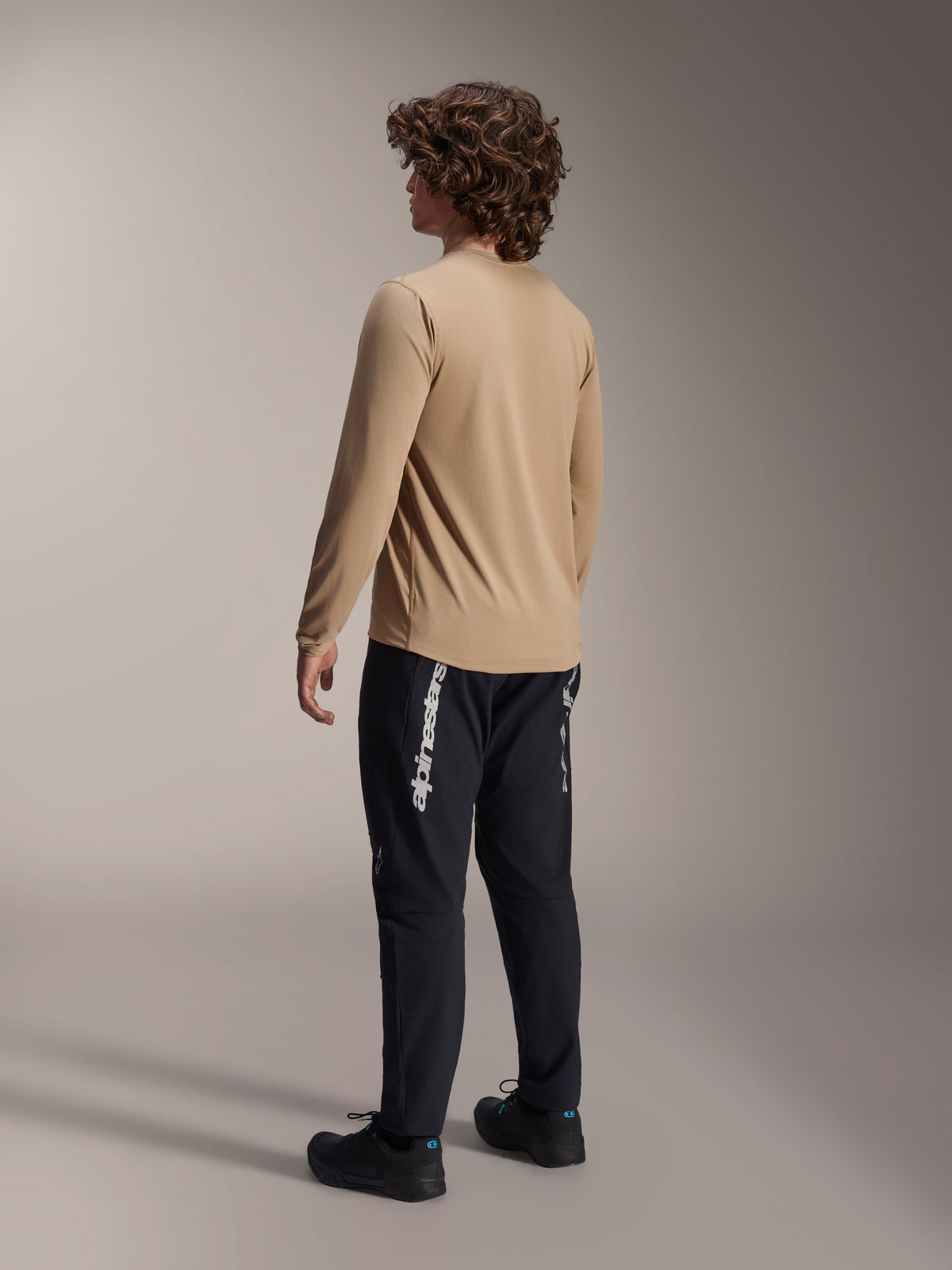 Alpinestars A-Dura Astar Long Sleeve Jersey in Light Brown, back view of a male model showcasing the drop tail design, paired with black pants and shoes, casual mountain bike performance gear.
