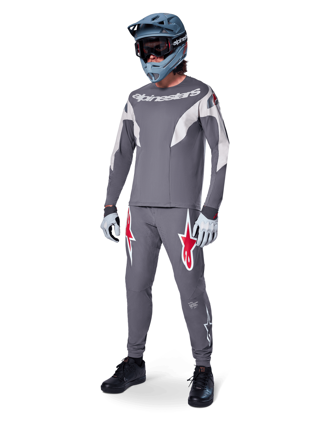 MTB rider wearing Alpinestars A-Supra Race Jersey, Long Sleeve, Dark Gray with light gray accents, matching dark gray pants featuring red star logo, gray full-face helmet with goggles, light gray gloves, coordinated outfit for mountain bike racing