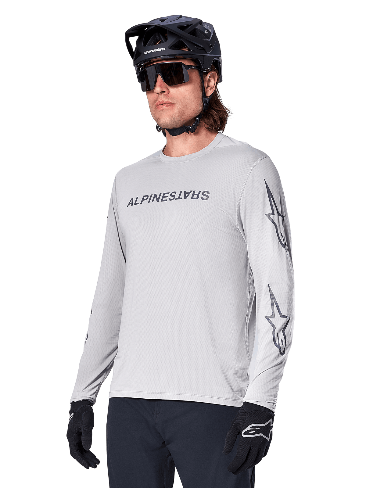 Alpinestars A-Dura Switch Langarmtrikot, Mountainbike-Trikot, Hellgrau, schwarzer Wortmarken-Siebdruck auf der Brust und Stern-Logos auf den Ärmeln, getragen mit schwarzem Mountainbike-Helm, dunkler Sonnenbrille und schwarzen Handschuhen, sportliche Offroad-Radfahrausrüstung