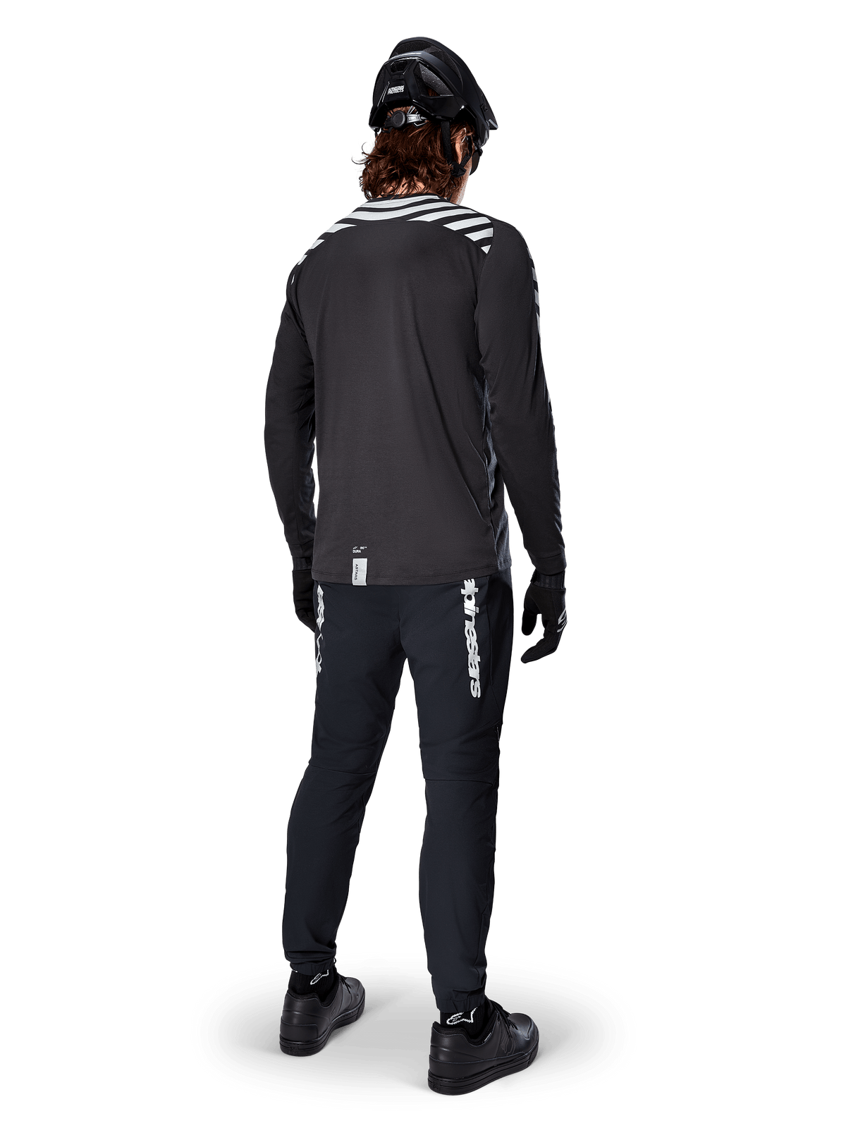 Mountainbike-Fahrer im Alpinestars A-Dura Dri Oscar Langarmtrikot in Black Reflective, Rückansicht, kombiniert mit schwarzer technischer Hose, Helm und Handschuhen, mit reflektierenden Streifengrafiken auf den Schultern und einem verlängerten Rücken für den Offroad-Radsport.