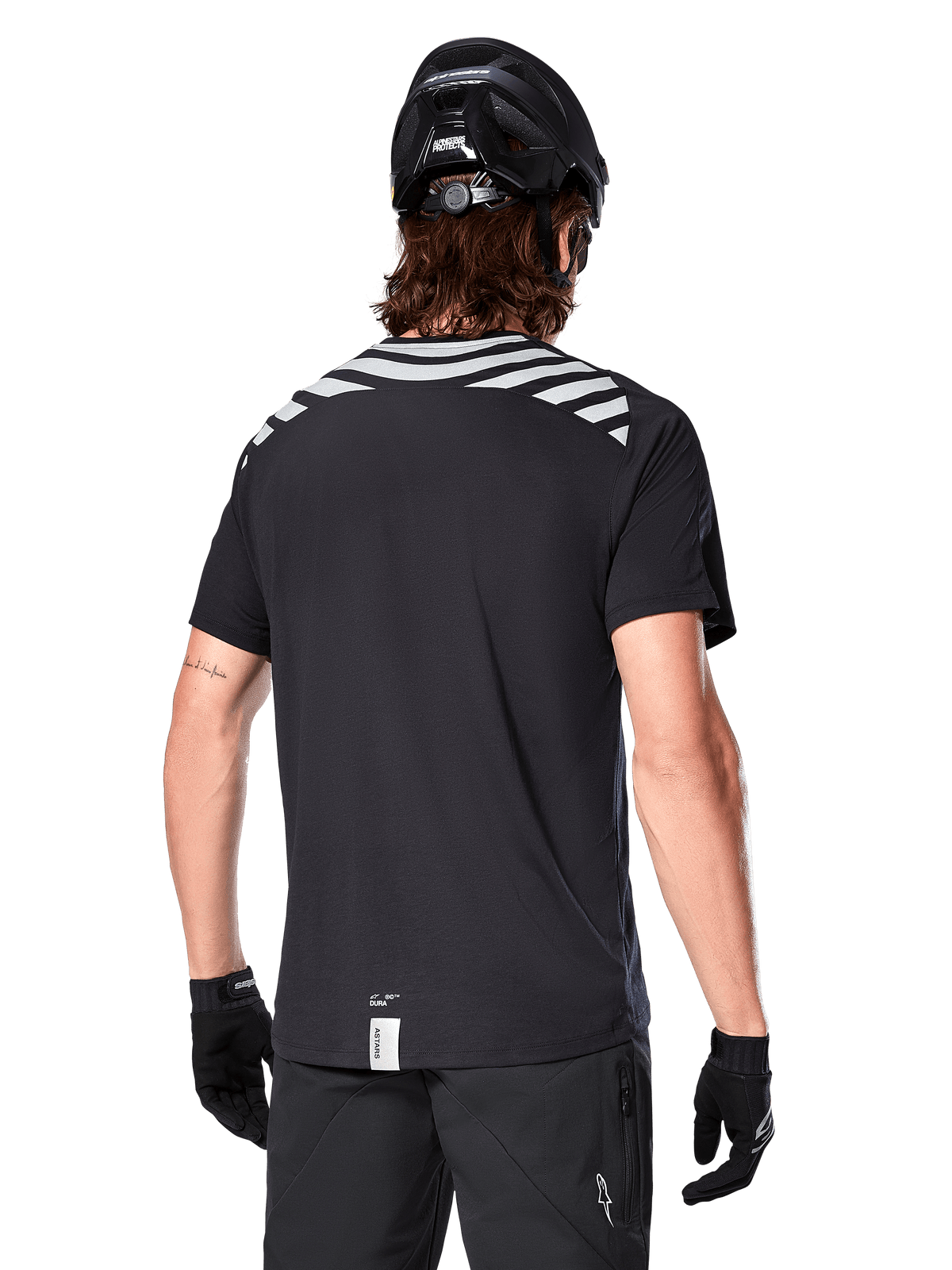 Alpinestars A-Dura Dri Oscar Trikot, Kurzarm-MTB-Trikot, Schwarz Reflektierend, Rückansicht am Model mit Fahrradhelm und Handschuhen, mit reflektierenden grauen Schulterstreifen und verlängertem Rückenteil, feuchtigkeitsableitendes Funktionsgewebe für Mountainbiking