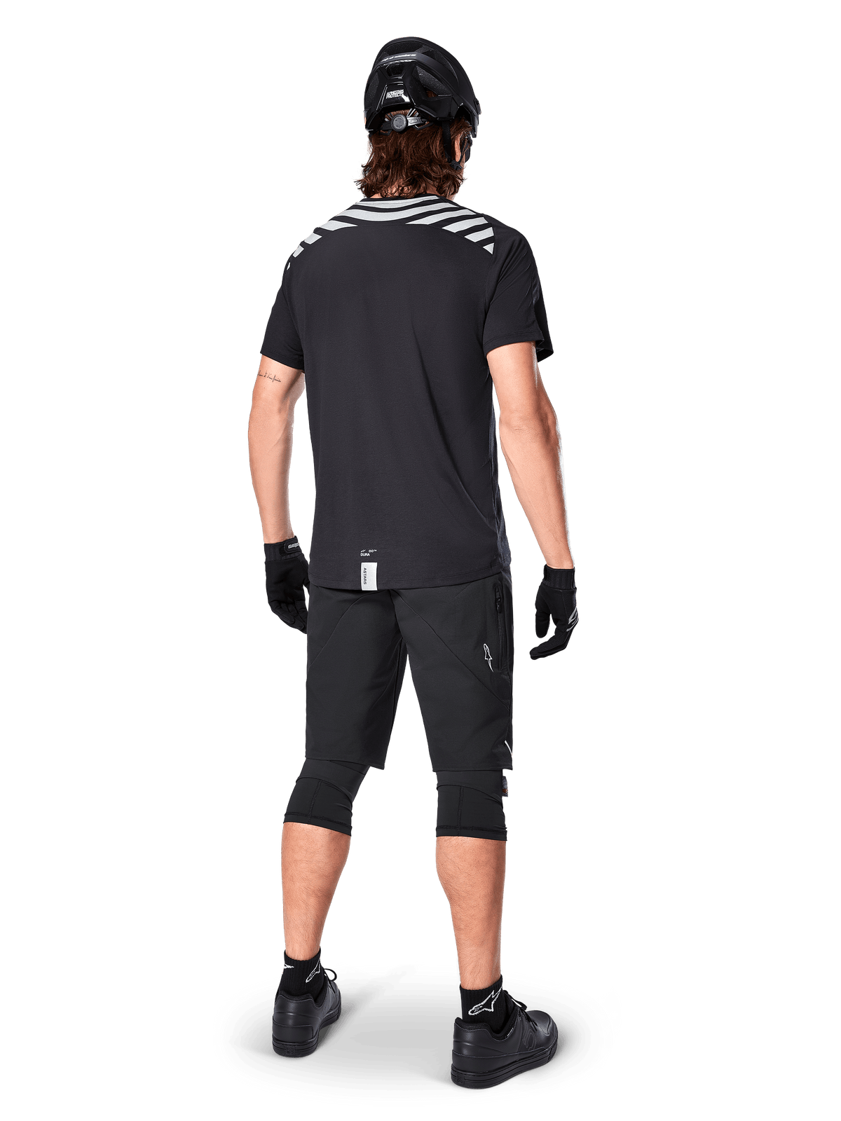 Alpinestars A-Dura Dri Oscar Kurzarmtrikot, Black Reflective, Rückansicht eines Fahrers mit schwarzen Mountainbike-Shorts, Helm und Knieschonern, reflektierende Schultergrafiken und verlängerte Rückenpartie für MTB-Fahrten.