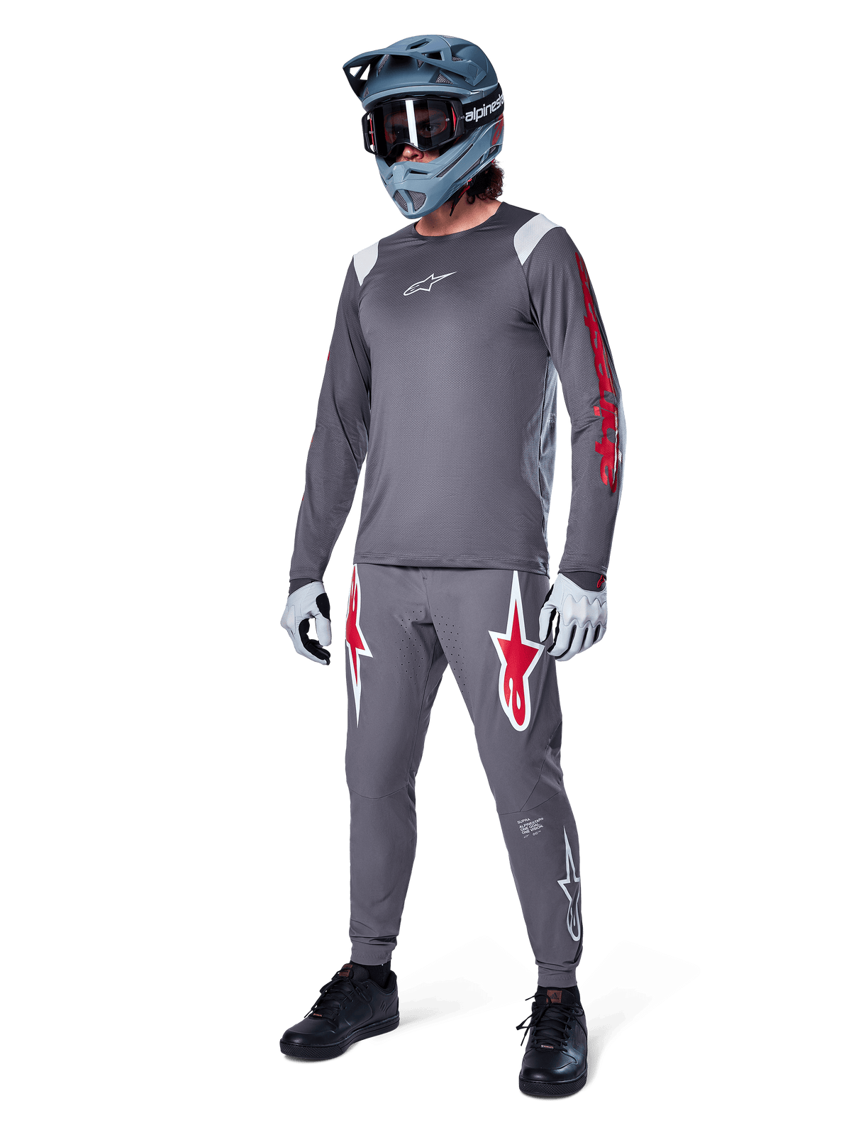 Alpinestars A-Supra Astar Long Sleeve Jersey in Dark Gray, featuring red branding on the sleeves and a white logo on the chest, worn by a rider with matching gray pants, a blue-gray full-face helmet, and performance gloves, coordinated mountain bike gear for gravity riding.