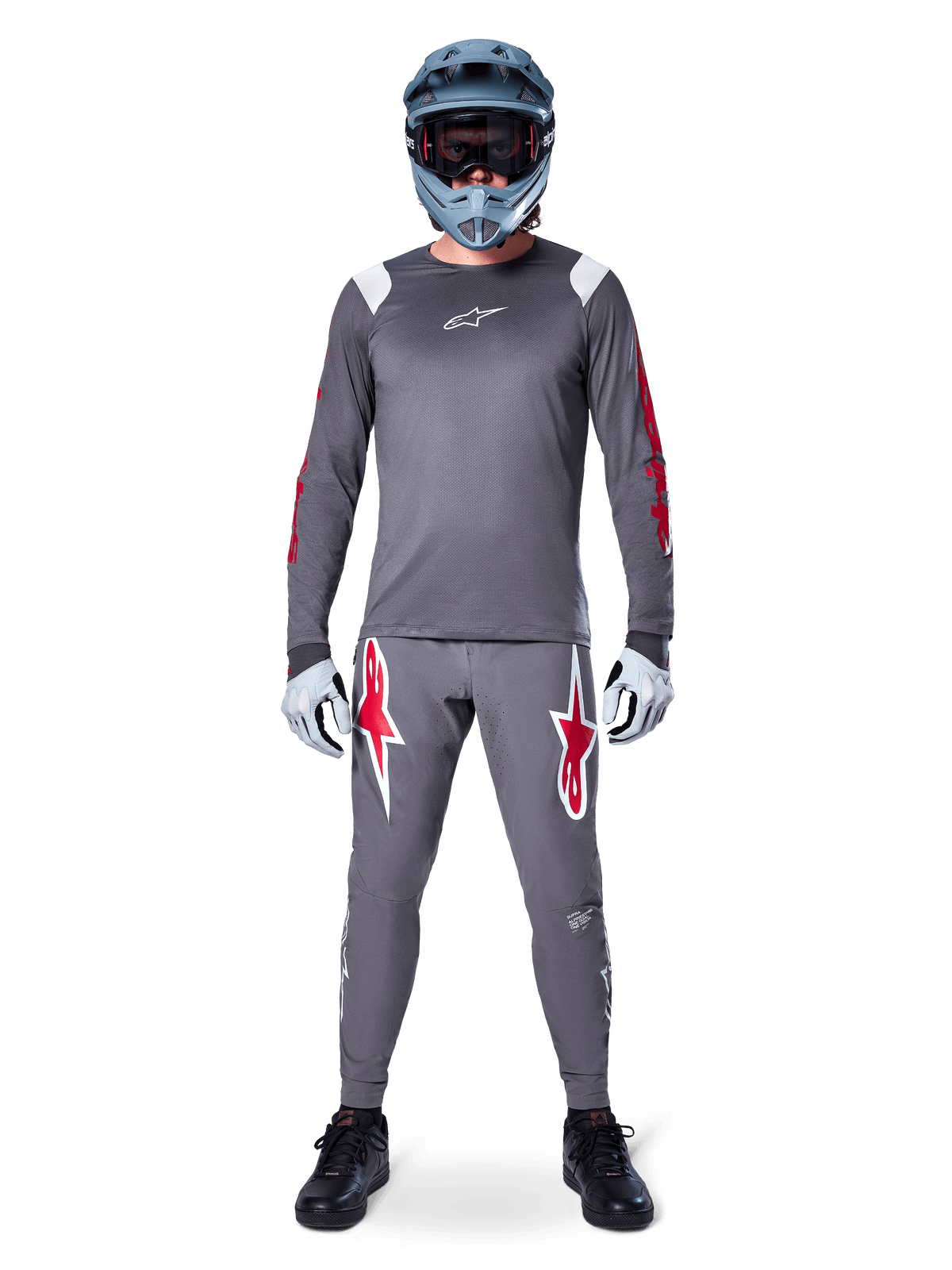 MTB rider wearing Alpinestars A-Supra Astar Long Sleeve Jersey, Dark Gray with red accents and white chest logo, matching gray pants with large red star graphics, blue full-face helmet with goggles, light gray gloves, and black shoes, professional mountain bike gear ensemble