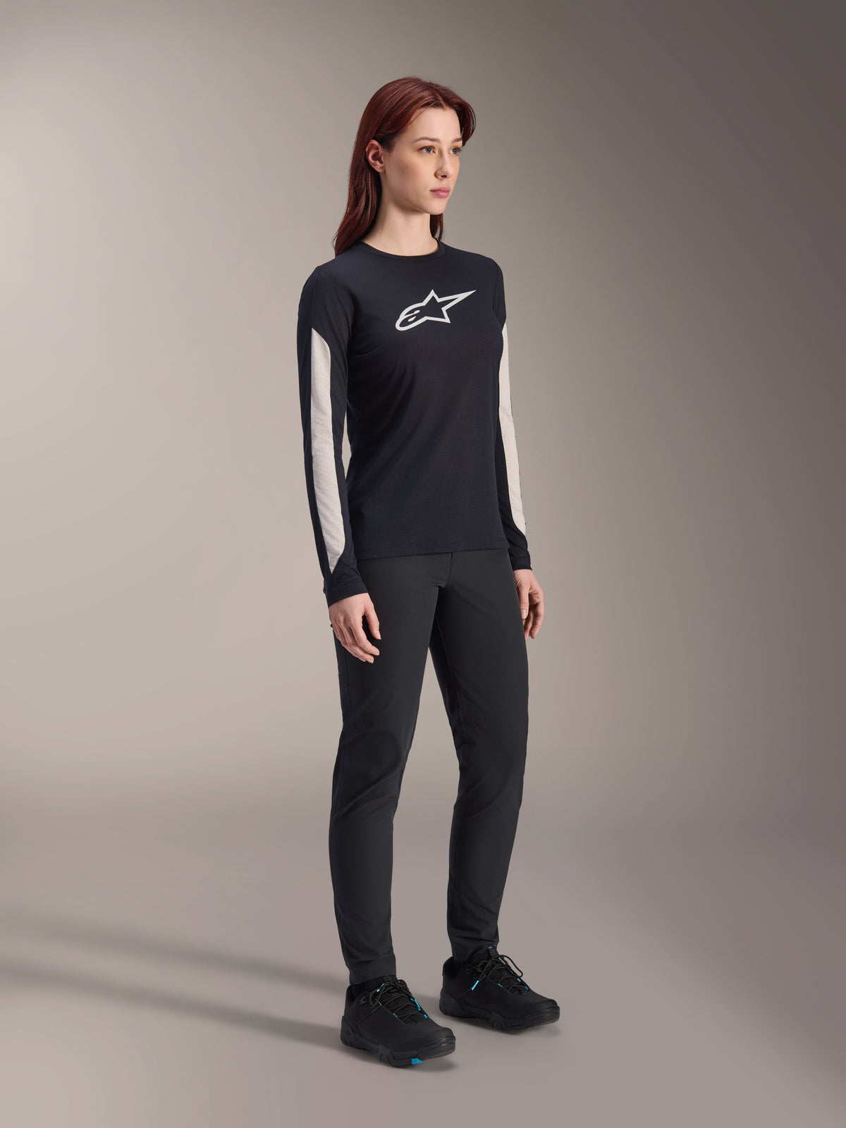 Woman wearing Alpinestars MTB gear, black Stella A-Dura Elite Jersey with white chest logo and sleeve panels, black Stella A-Dura Pants, black cycling shoes with blue accents, full performance outfit for mountain biking