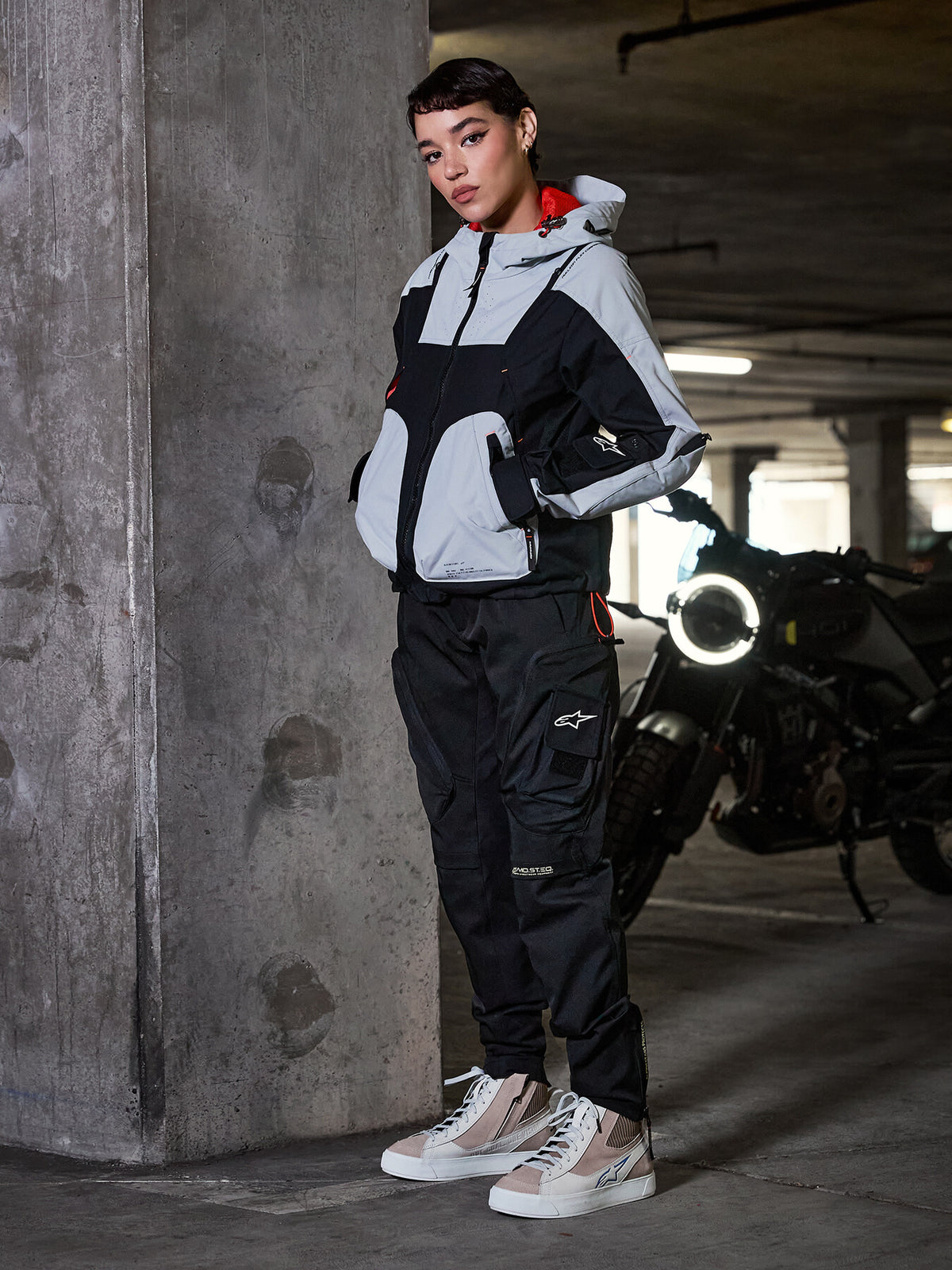 Alpinestars Actea Damen-Hose, Schwarz, Motorradfahrerin in einer Motorradhose im Cargo-Stil, grau-schwarze technische Jacke und beige Sneakers, urbane Parkhaus-Umgebung mit Motorrad