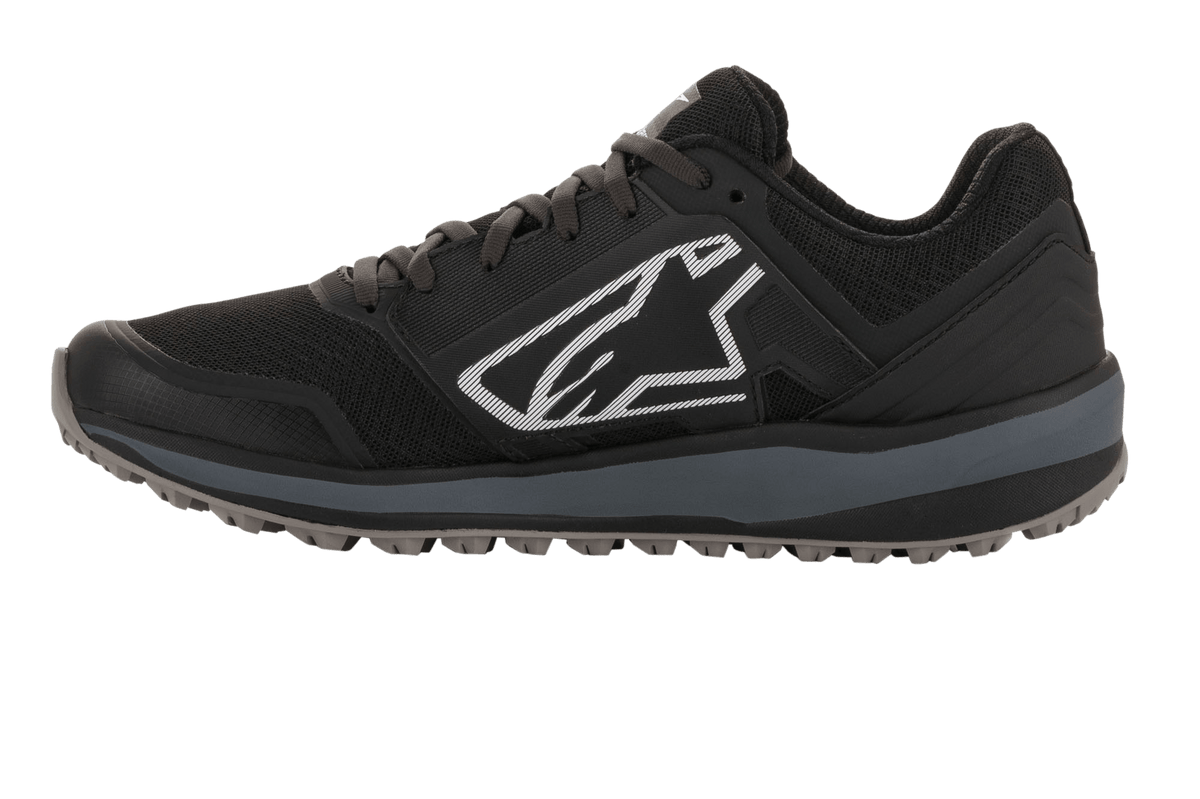 Alpinestars Meta Trail Schuhe, Performance-Laufschuh, Schwarz und Dunkelgrau, Seitenprofil mit großem weißen Astars-Logo, dunkelgraue Zwischensohle, profilierte ölbeständige Gummisohle, konzipiert für Komfort im Fahrerlager und Motorsport-Crews.
