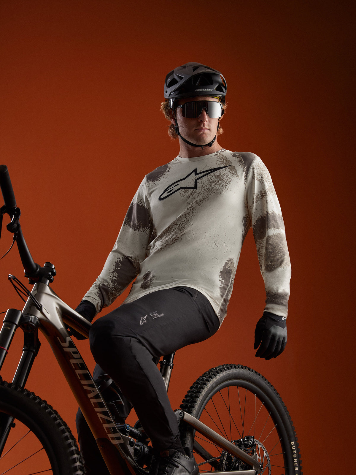 A-Dura Ride Langarmtrikot in Sand Camo mit Alpinestars-Logo, getragen von einem MTB-Fahrer auf einem Mountainbike.