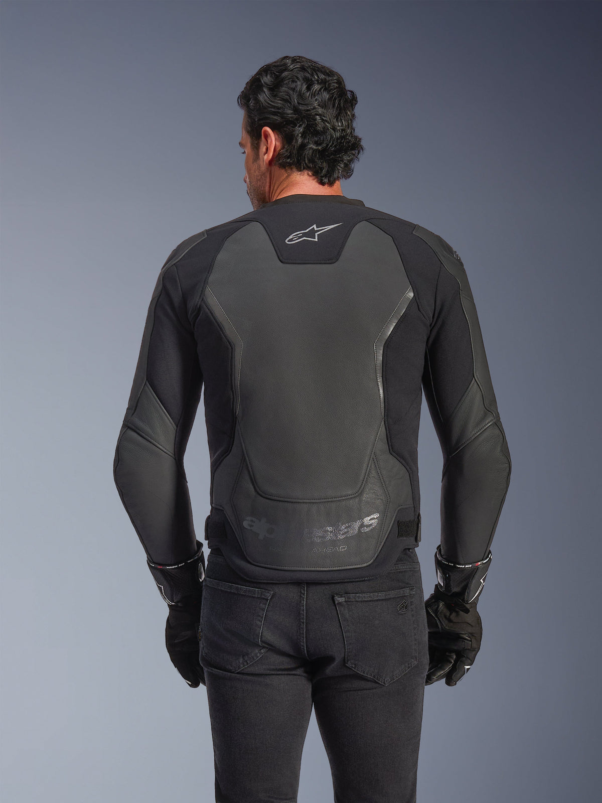 Alpinestars GP Force V2 Lederjacke, Schwarz, Rückansicht eines Mannes, der die Jacke mit schwarzen Handschuhen und Jeans trägt, mit großen Leder-Schutzpaneelen am Rücken, Alpinestars-Logo am oberen Rücken und geprägtem Branding an der unteren Taille, sportlich inspirierte Motorradausrüstung
