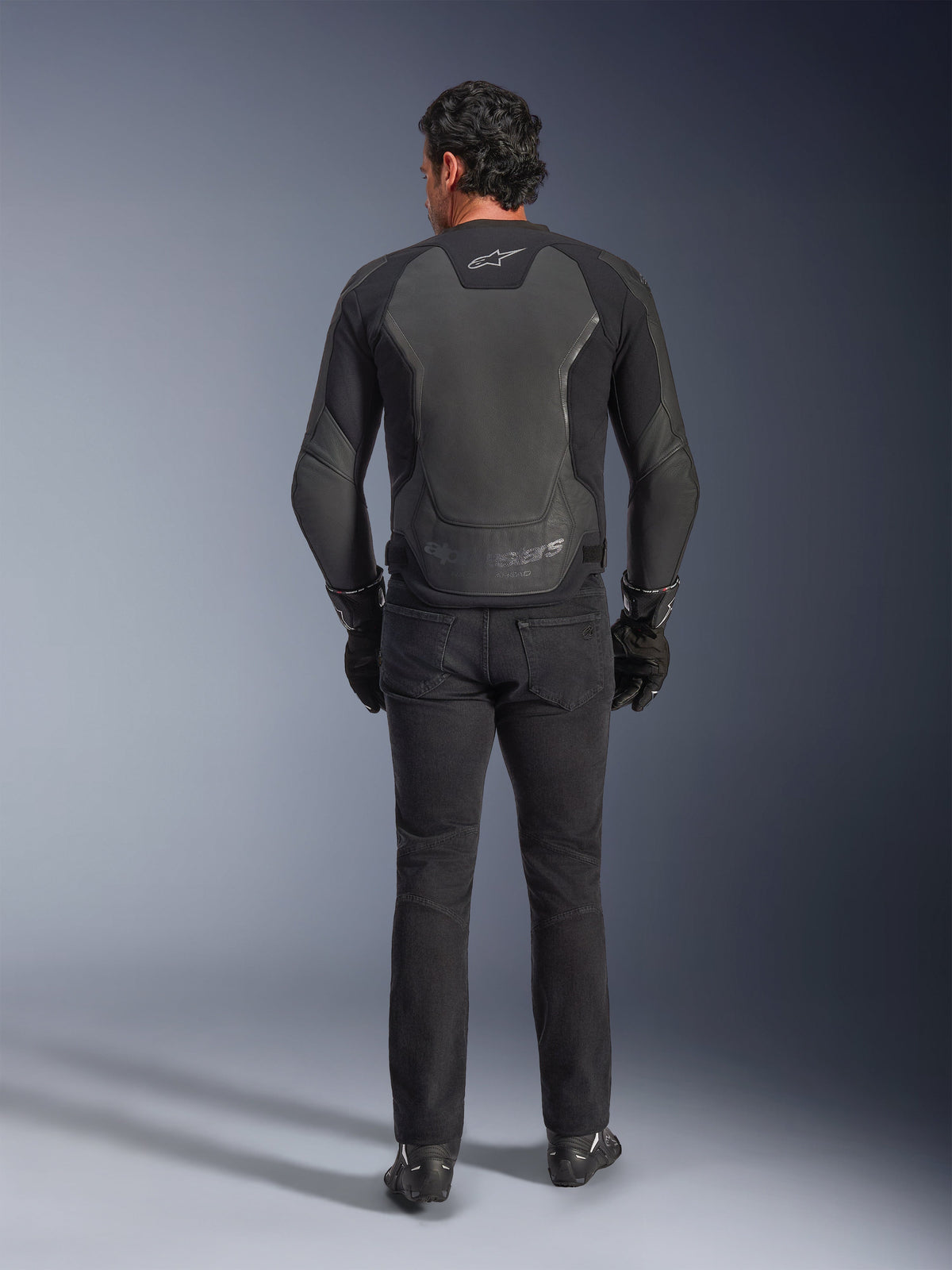 Alpinestars GP Force V2 Lederjacke, Schwarz, Rückansicht, hochwertige Sport-Motorradjacke mit Leder-Textil-Konstruktion, getragen mit schwarzen Motorradjeans und Handschuhen, mit schützenden Schulterpaneelen und Branding-Details