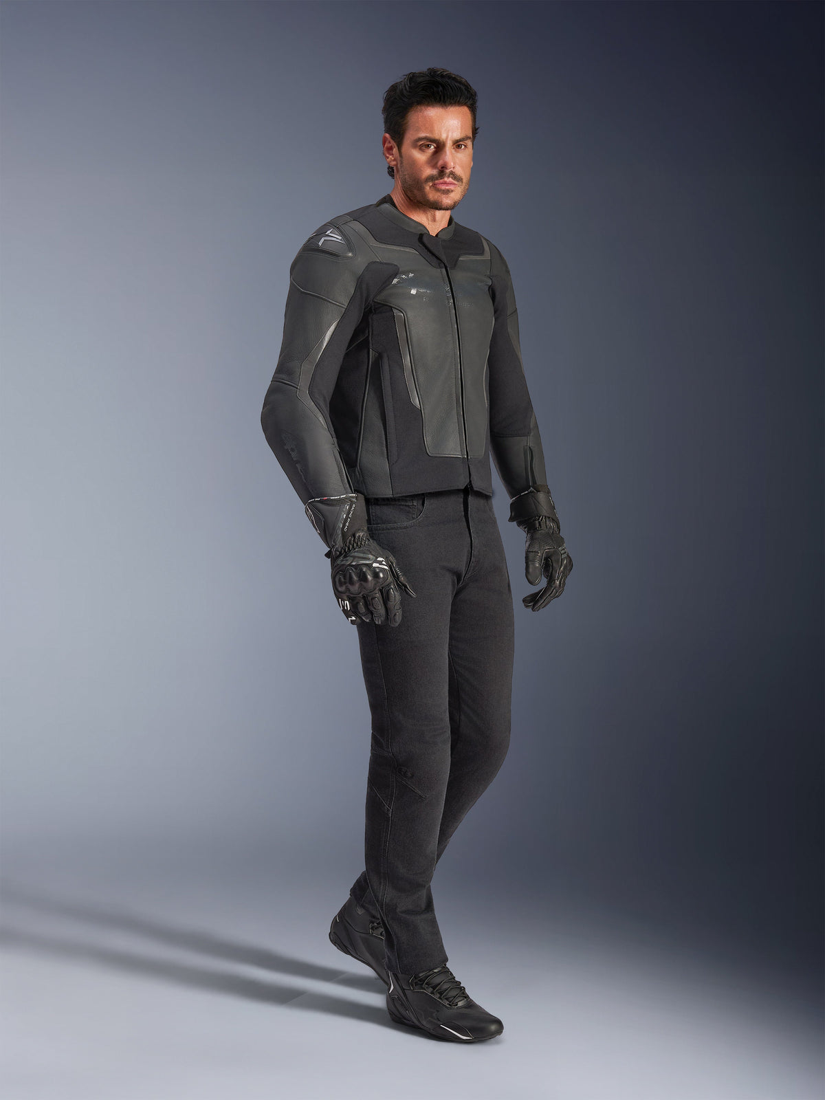 Mann trägt Alpinestars GP Force V2 Lederjacke in Schwarz, kombiniert mit schwarzer Hose und Motorradhandschuhen, Ganzkörper-Studioaufnahme, hochwertige Rindslederkonstruktion mit integriertem Schulter- und Ellbogenschutz, professionelle Sport-Motorradbekleidung