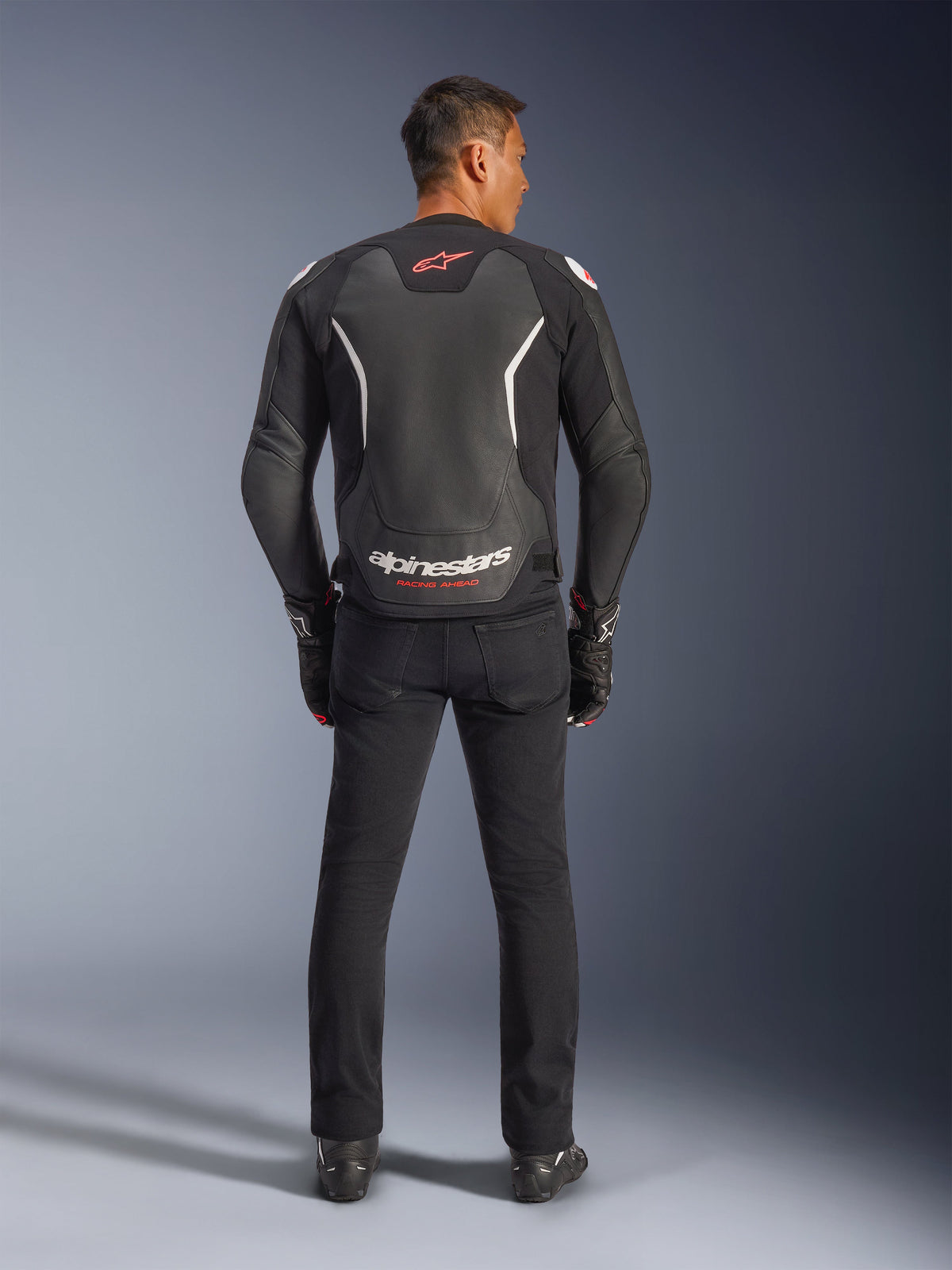 Alpinestars GP Force V2 Lederjacke, Schwarz und Weiß, Rückansicht, getragen von einem Fahrer mit schwarzer Hose und Handschuhen, großes weißes Branding am unteren Rücken und rote Akzente, hochwertige Rindslederkonstruktion für Motorrad-Straßenausrüstung