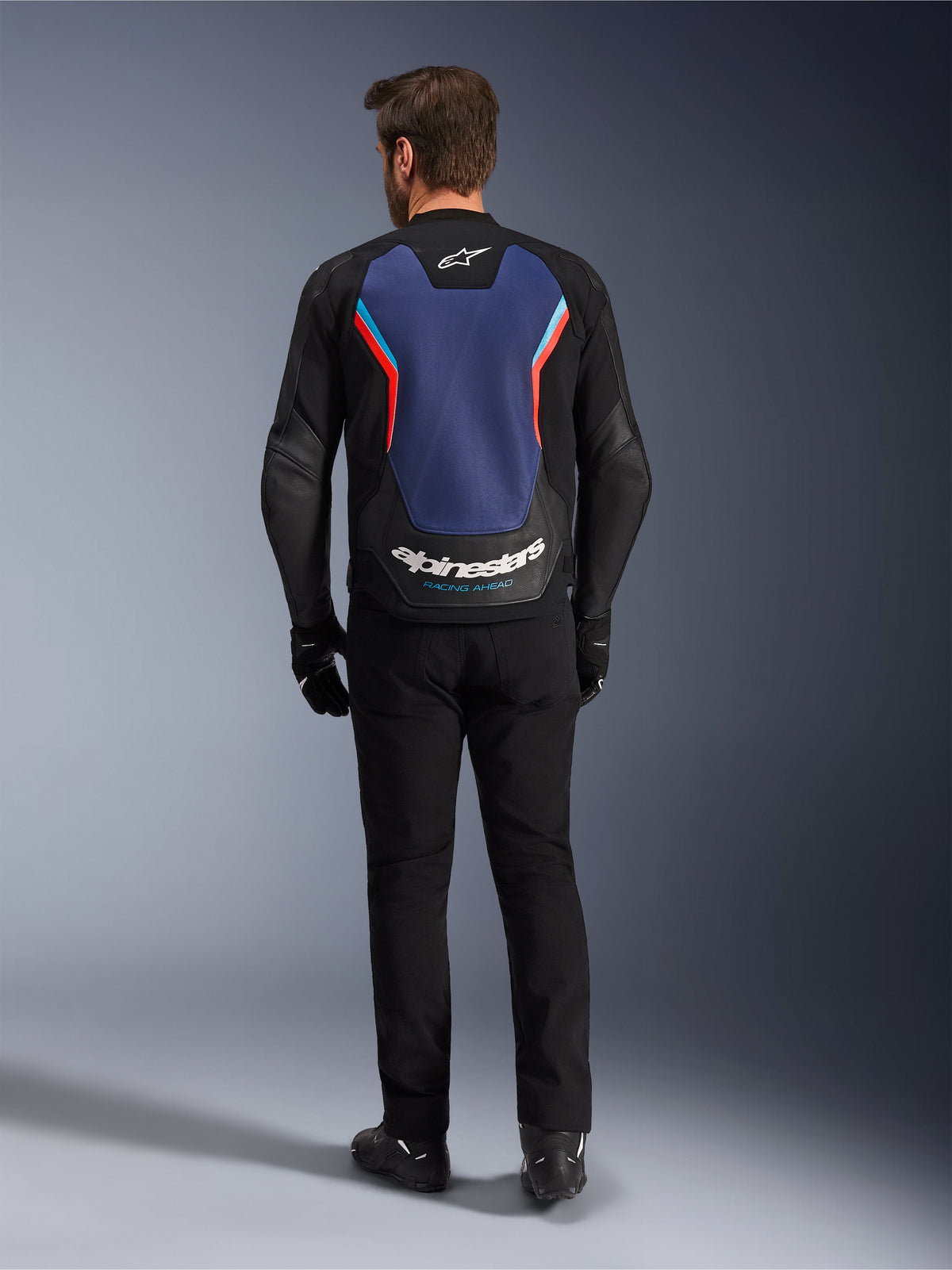 Alpinestars GP Force V2 Lederjacke, Sport-Motorradjacke, Schwarz Blau Rot Fluo, Rückansicht an einem Model, mit großem blauem Rückenteil, roten und hellblauen Akzenten sowie weißem Alpinestars-Branding am unteren Rücken, kombiniert mit schwarzer Motorradhose und Handschuhen.