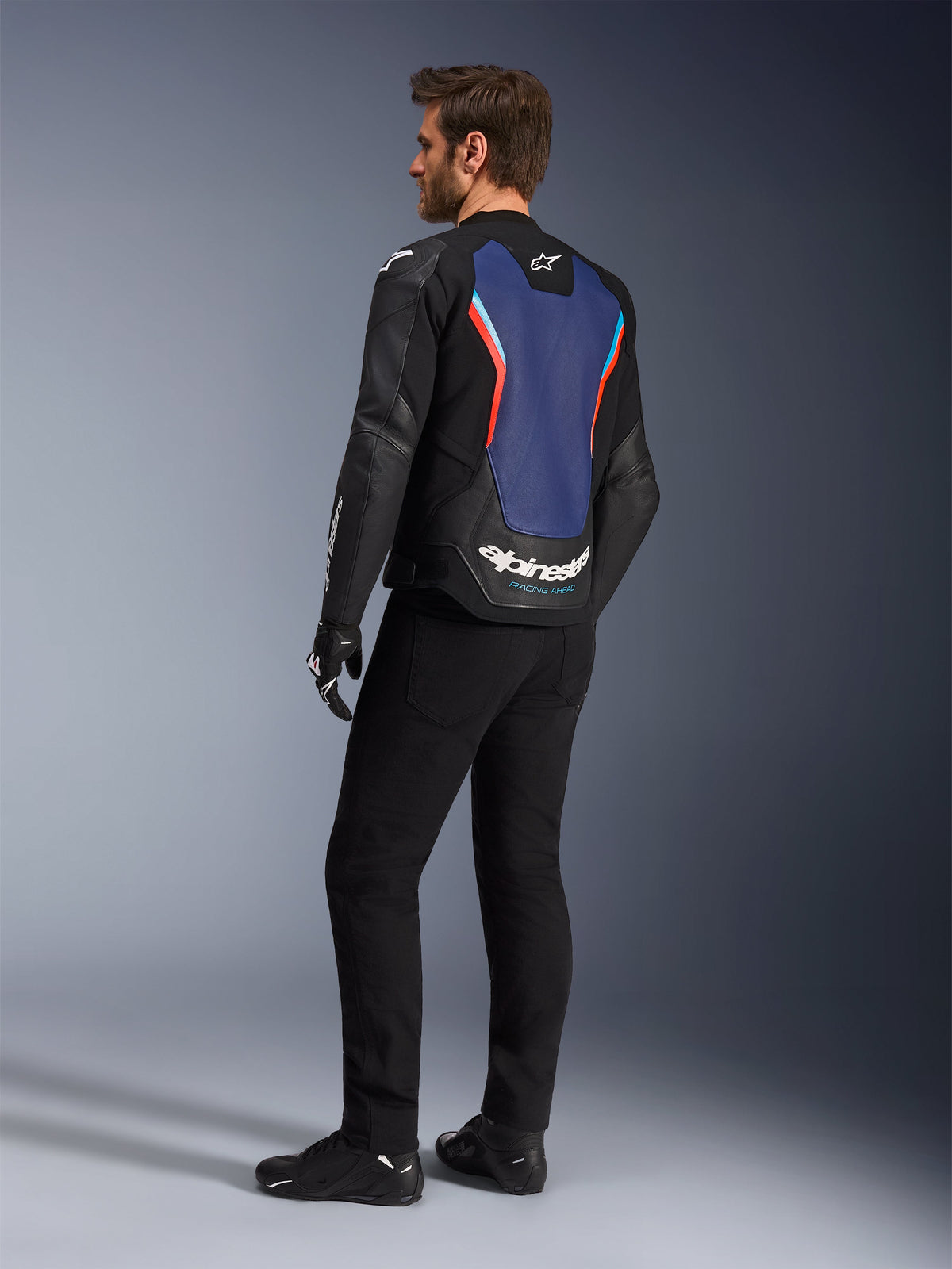 Dreiviertel-Rückansicht eines männlichen Models in der Alpinestars GP Force V2 Lederjacke in Schwarz Blau Rot Fluo, die das blaue Rückenpanel mit roten fluoreszierenden Akzenten und weißem Branding hervorhebt, kombiniert mit einer schwarzen Hose und Motorradhandschuhen.