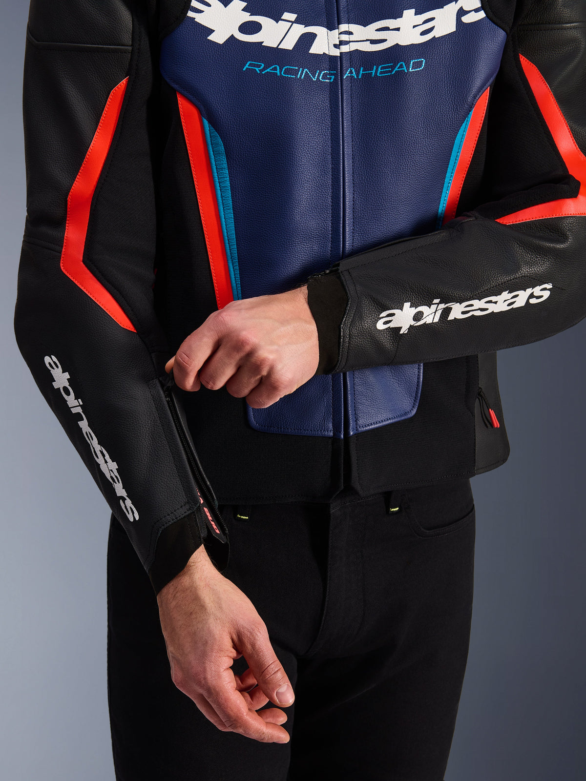 Alpinestars GP Force V2 Lederjacke in Schwarz, Blau und Fluo-Rot, Nahaufnahme einer Person beim Einstellen des Reißverschlusses am Ärmelbündchen, weiße Alpinestars-Logos auf Unterarm und Brust, strukturierte Rindslederkonstruktion für das sportliche Motorradfahren