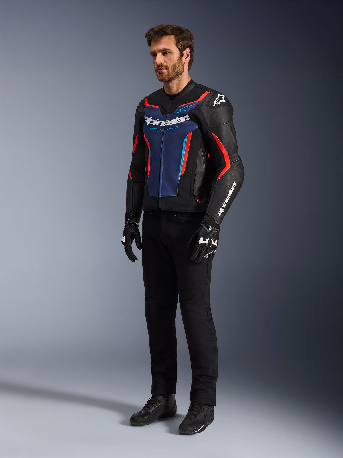 Model trägt die Alpinestars GP Force V2 Lederjacke in Schwarz Blau Rot Fluo, kombiniert mit schwarzer technischer Fahrhose, schwarzen Handschuhen und Stiefeln, professionelle Motorrad-Rennausrüstung für sportliches Fahren
