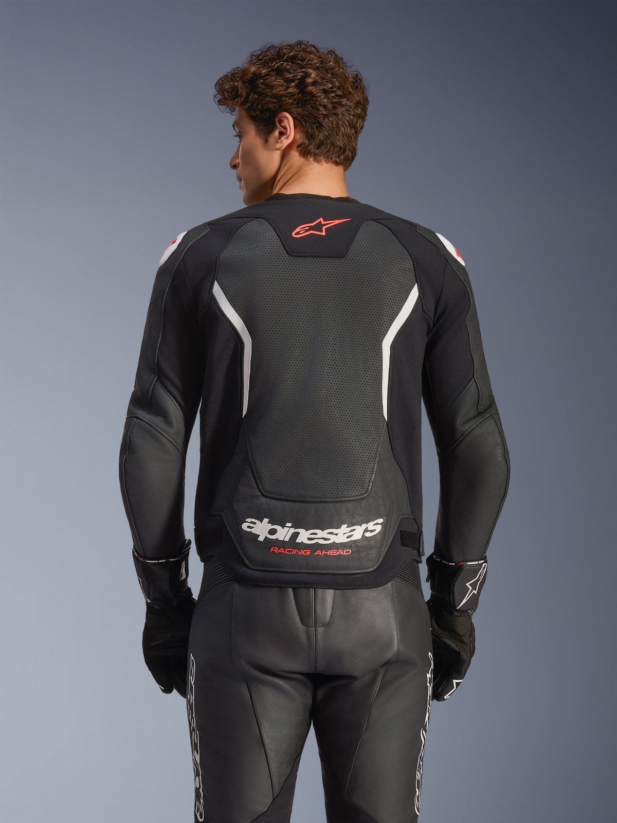 Alpinestars GP Force V2 Air Lederjacke, Schwarz mit weißen und roten Akzenten, Rückansicht mit perforiertem Leder-Rückenpanel, weißes Alpinestars-Branding und rote Logo-Details, getragen mit passender schwarzer Leder-Rennhose und Handschuhen.