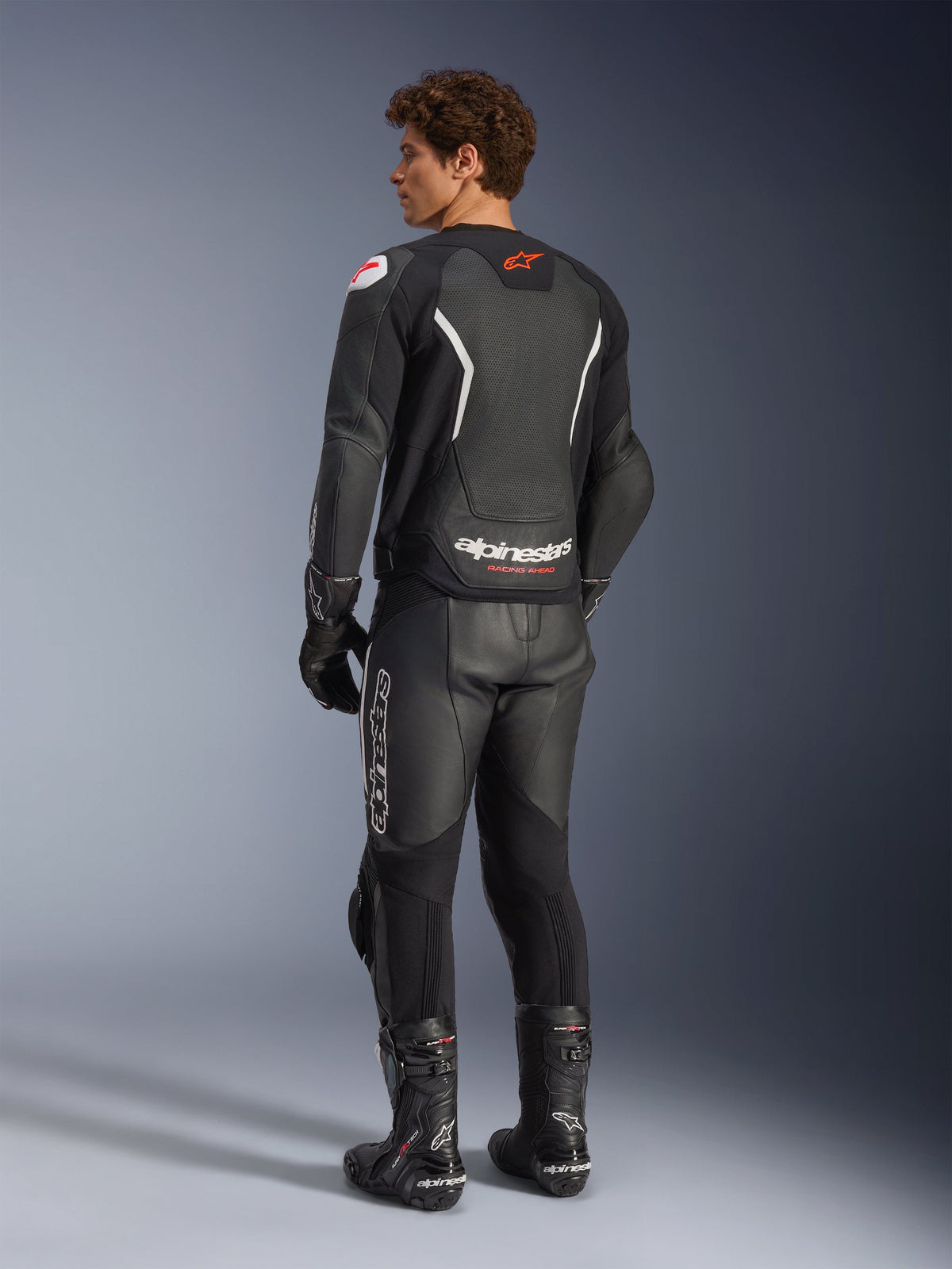 Alpinestars GP Force V2 Air Lederjacke, Schwarz und Weiß, Rückansicht am Modell mit perforiertem Rückenteil zur Belüftung, weißem Branding und rotem Stern-Logo, getragen mit passender Leder-Rennhose, Handschuhen und Stiefeln