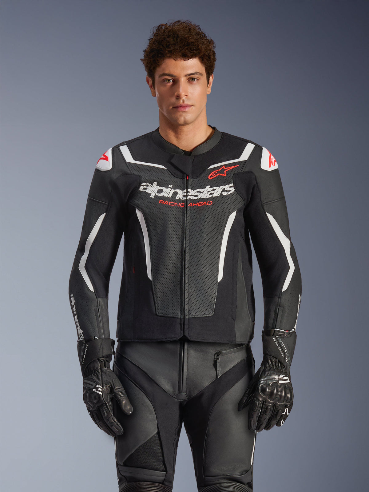 Alpinestars GP Force V2 Air Lederjacke, Schwarz mit weißen und roten Akzenten, Model trägt eine perforierte Lederjacke mit „alpinestars“- und „RACING AHEAD“-Branding auf der Brust, kombiniert mit passender Rennhose und Handschuhen, Premium-Sport-Motorradausrüstung