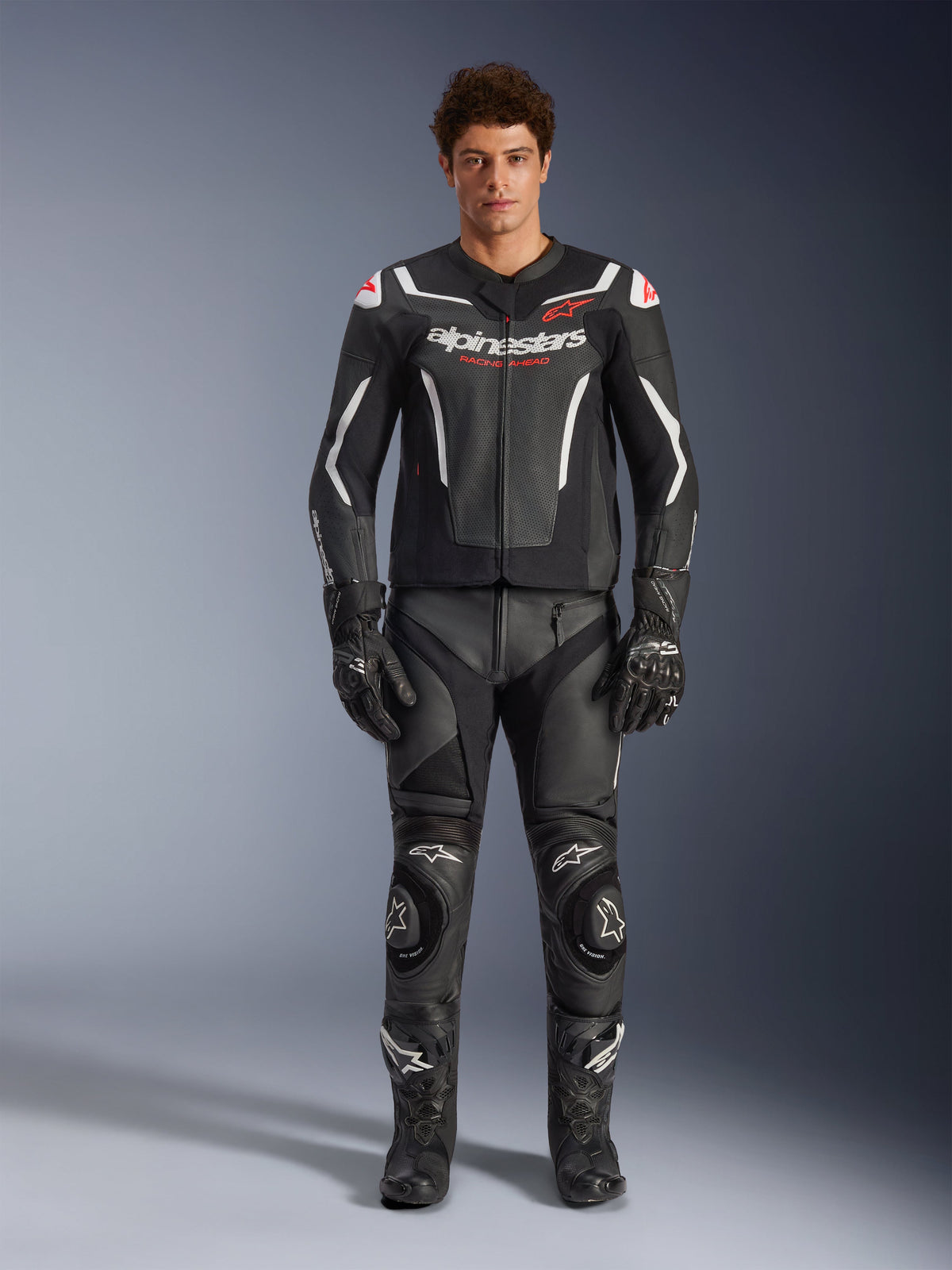 Männliches Model trägt Alpinestars GP Force V2 Air Lederjacke in Schwarz und Weiß, kombiniert mit passender Lederhose, Handschuhen und Stiefeln, sportliche Frontansicht, perforierte Lederkonstruktion mit weißen Akzenten und Alpinestars-Logos