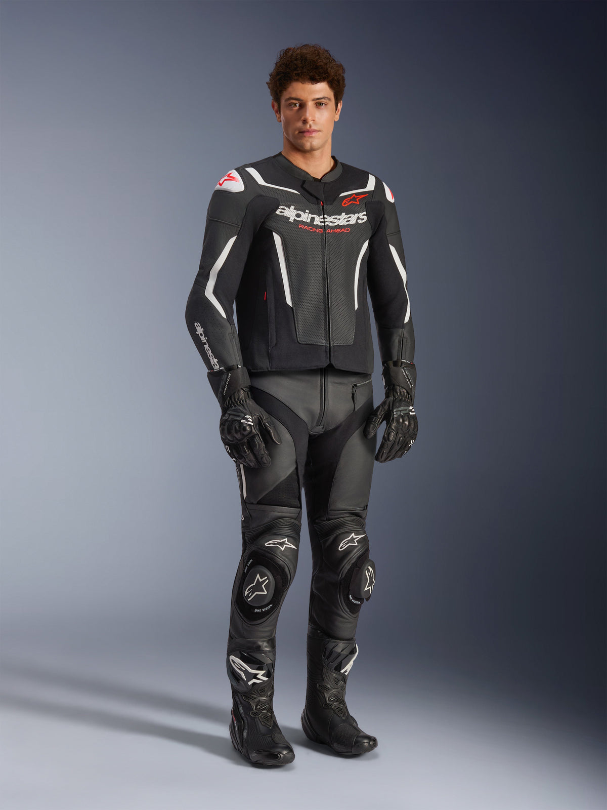 Alpinestars GP Force V2 Air Lederjacke in Schwarz und Weiß, getragen von einem männlichen Fahrer mit passender schwarzer Rennhose, Handschuhen und Stiefeln, mit perforierten Ledereinsätzen, weißen Akzenten und rotem Alpinestars-Logo auf der Brust, Sportmotorrad-Ausrüstung