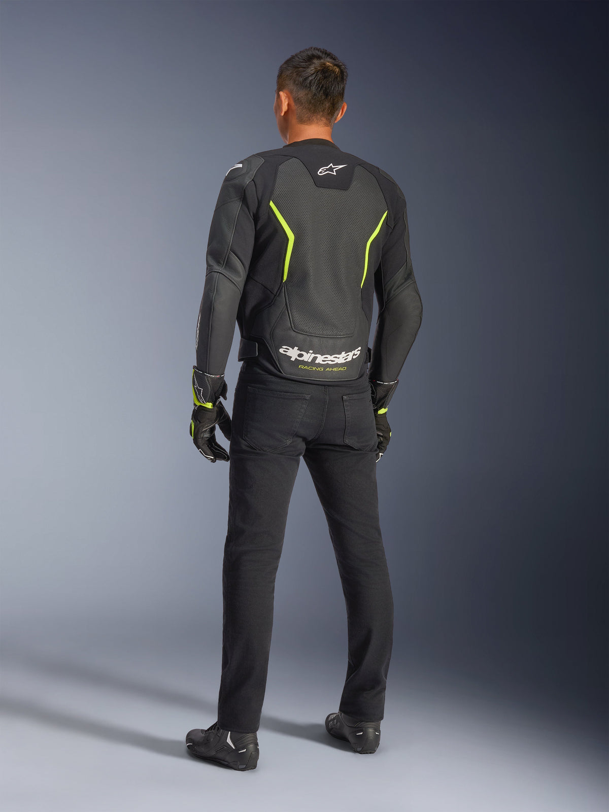 Alpinestars GP Force V2 Air Lederjacke, Schwarz und Gelb Fluo, Rückansicht eines Fahrers in der perforierten Lederjacke, markante gelbe Akzente, weißes Alpinestars-Logo mit „RACING AHEAD“ am unteren Rücken, kombiniert mit schwarzer Motorradhose und Stiefeln.
