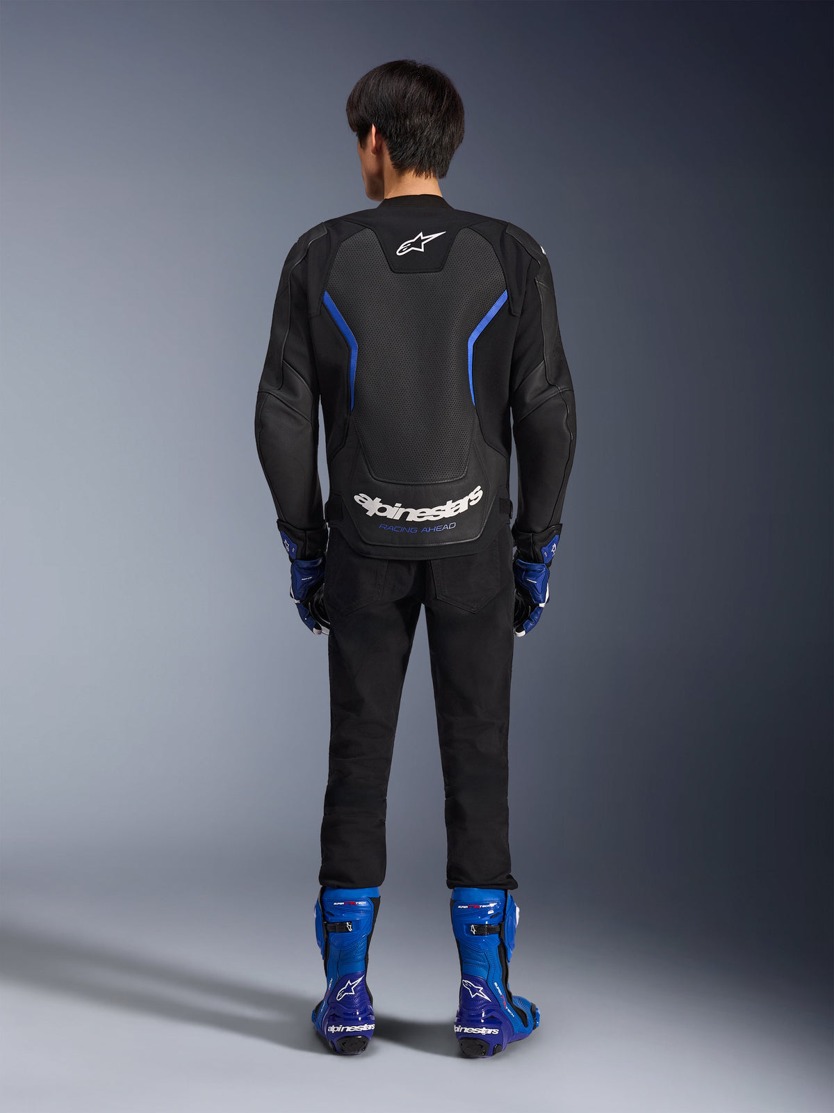 Alpinestars GP Force V2 Airflow Leather Jacket, Black Metallic Blue, rear view showing perforated leather panels and white branding, worn with blue racing boots and black pants, sport motorcycle racing gear